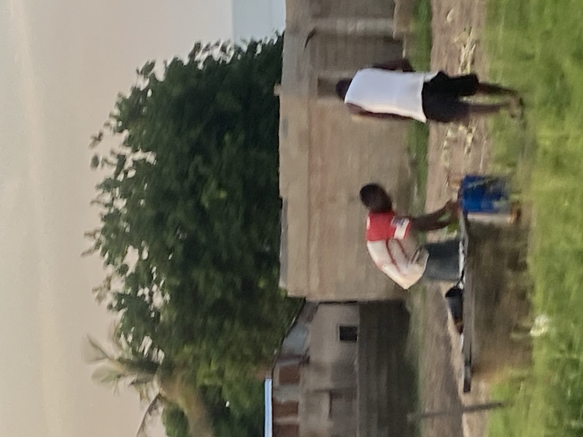 In Jauro Yinu, Nigeria, two individuals are observed outdoors amidst a sparsely vegetated environment with a cloudy sky. In the foreground, a person wearing a red and white shirt is bending over a concrete structure, appearing to interact with objects. To their right, another individual in a white tank top and black shorts stands facing away from the viewer. Behind them, unfinished or partially constructed buildings are visible. Lush green trees form a backdrop, with a hint of the ocean in the distance.