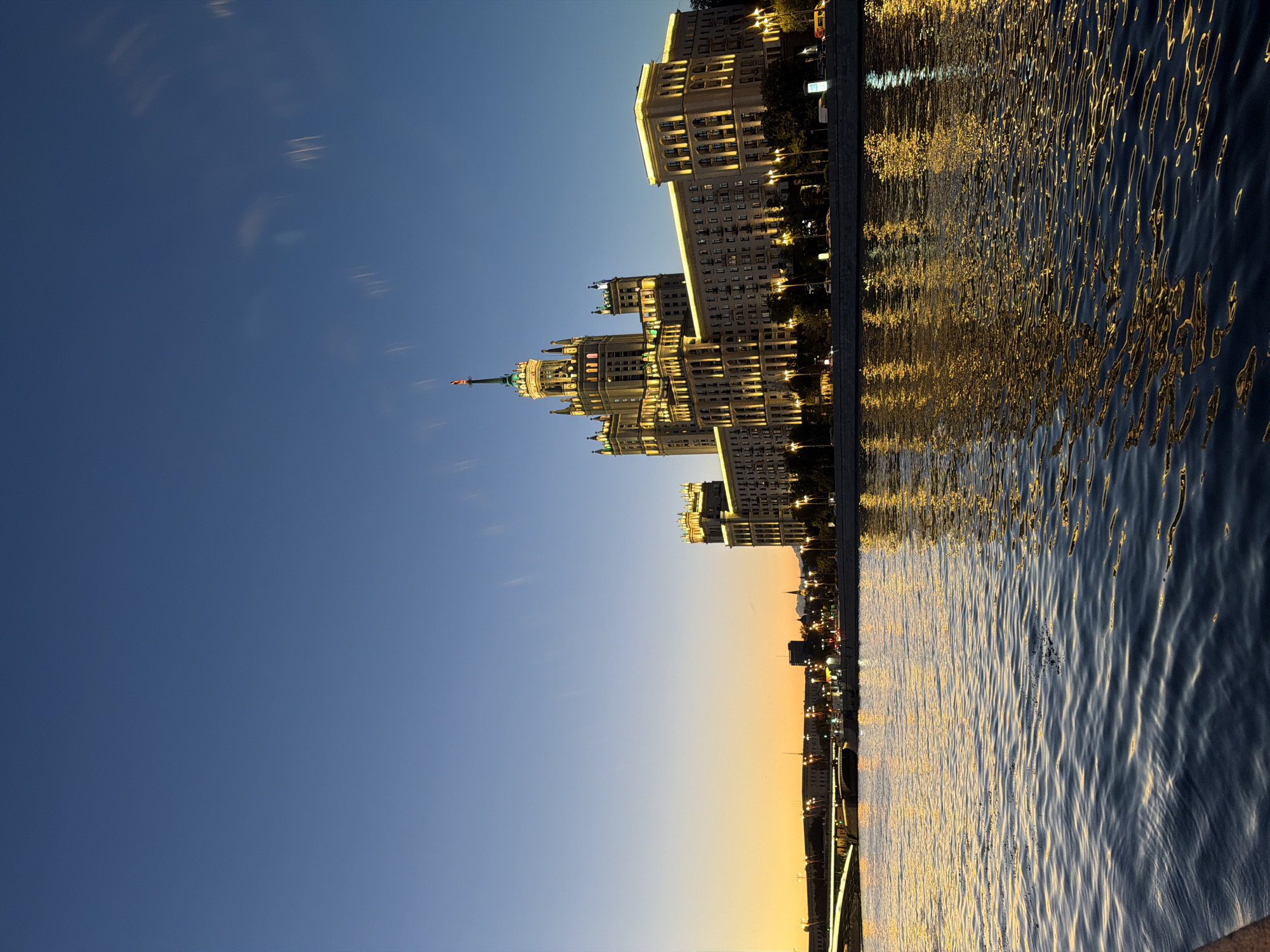 The image captures a twilight view of the Kotelnicheskaya Embankment Building in Moscow, Russia, situated along the Moskva River. The sky transitions from a deep blue at the top to a warm orange glow near the horizon, indicating the lingering light of sunset or the approaching dawn. The imposing Stalinist skyscraper, illuminated by numerous lights, stands prominently on the right side of the frame. Its distinctive tiered architecture, topped with a spire, is clearly visible.
Across the river, the embankment is lined with other buildings, also lit up, and a bridge is visible in the distance to the left, with streetlights creating a line of illumination. The Moskva River occupies the foreground and midground, its dark water reflecting the golden lights of the city, creating shimmering patterns. The water's surface shows subtle ripples. The overall scene depicts a city at dusk, with the architectural grandeur of Moscow illuminated against the darkening sky. There are no people or active interactions depicted in the image.