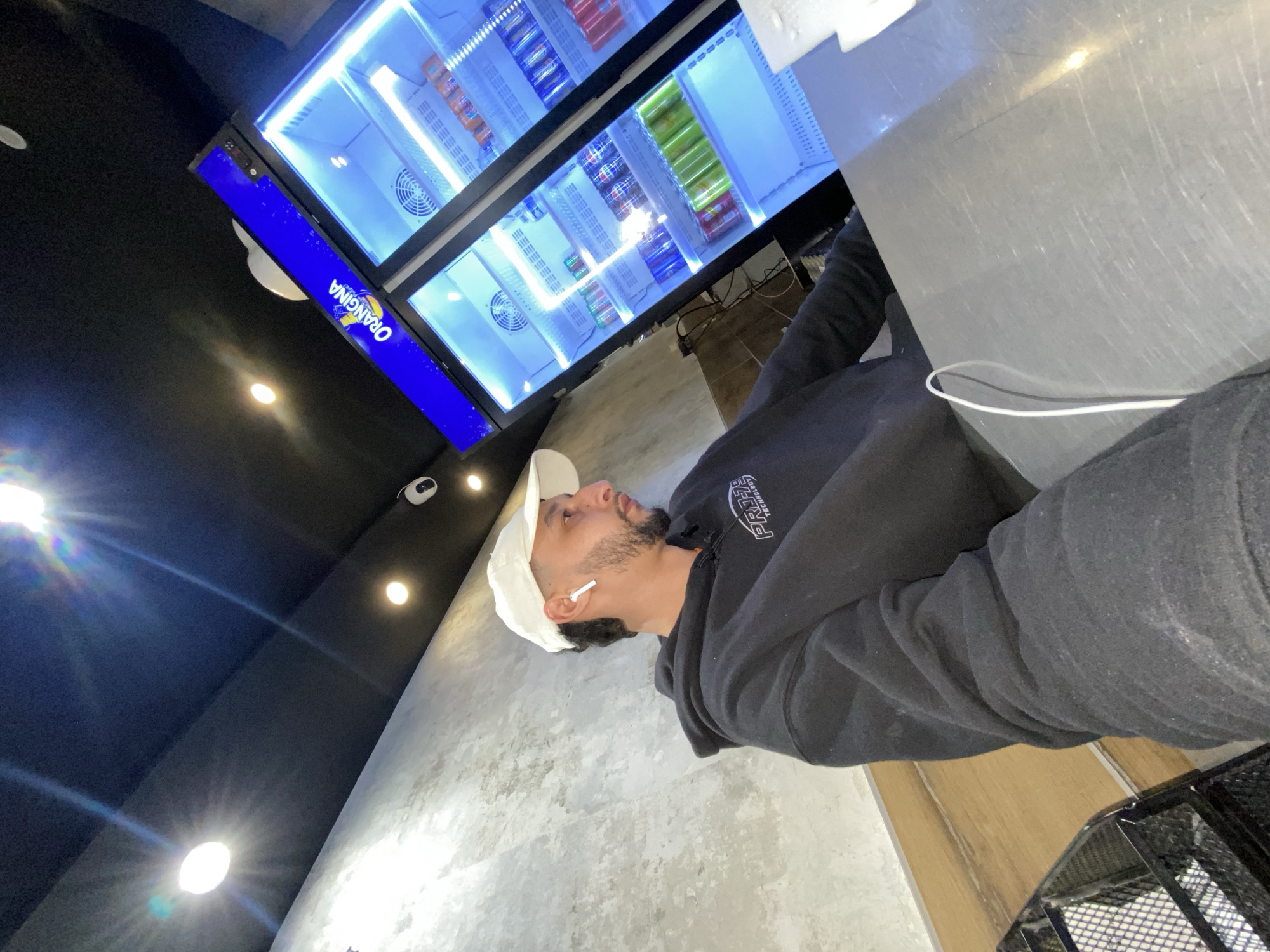 The image depicts a person standing indoors near a beverage refrigerator, presumably in Schiltigheim, France. The person is wearing a white baseball cap, wireless earbuds, and a black hooded sweatshirt. The refrigerator is stocked with various canned and bottled drinks, including Orangina. The interior walls are light-colored, and the ceiling is dark with recessed lighting. A wire basket is visible in the lower right corner of the image.