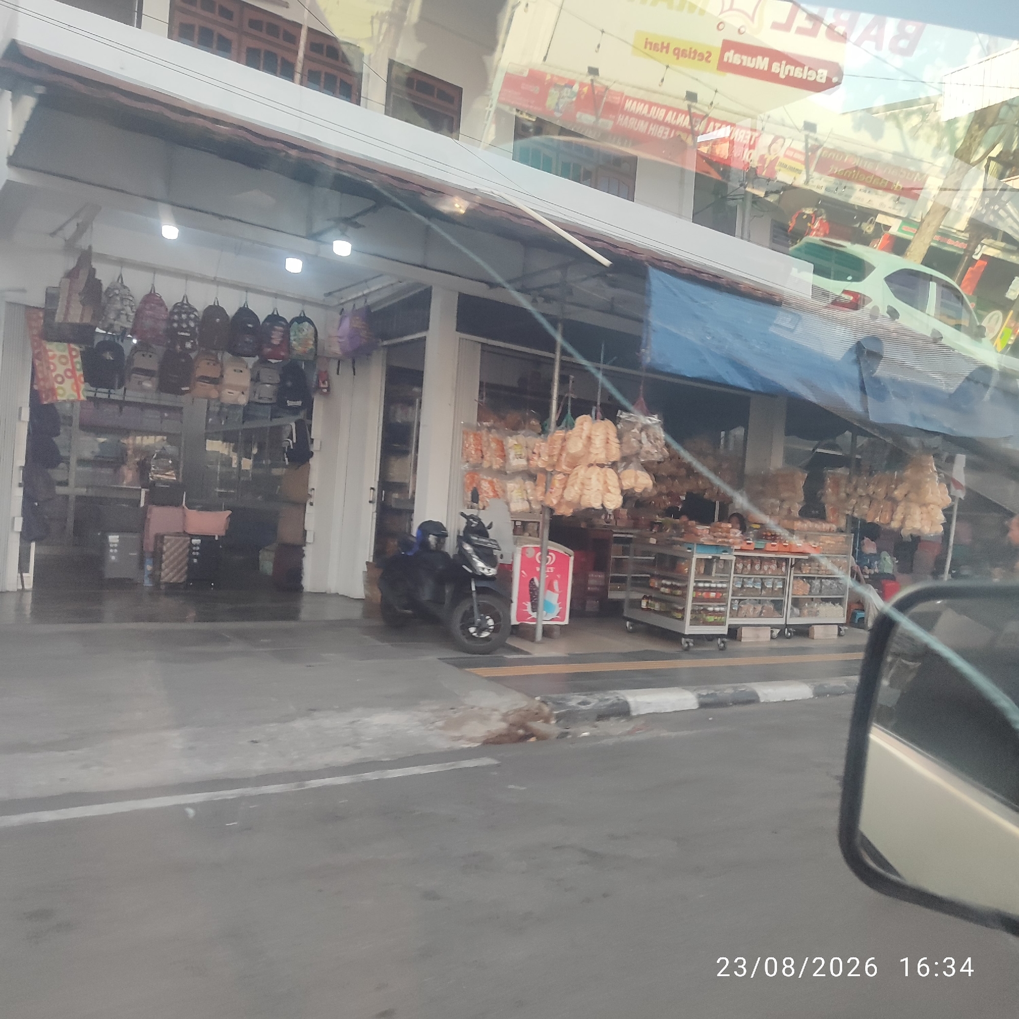 The image depicts a street scene in Tanjung Pandan, Indonesia, on August 23, 2026, at 4:34 PM. The foreground is dominated by the reflection of a car's side mirror and its interior, with the road and a sidewalk visible. On the sidewalk, there are scattered debris and a gutter.
The main subject of the image is a row of storefronts. One store displays numerous backpacks of various colors and designs hanging from the ceiling. Another store features shelves filled with packaged snacks and goods. A black motorcycle is parked on the sidewalk in front of one of the shops.
The storefronts themselves appear to be made of white and grey materials, with some featuring large glass windows. The upper levels of the buildings show traditional Indonesian architecture with wooden window frames. Reflections on the glass surfaces of the buildings reveal more of the street, including a light-colored car driving by and signage. One sign, partially obscured by reflections, appears to be in Indonesian and advertises "BabeL". The overall impression is that of a bustling commercial area.