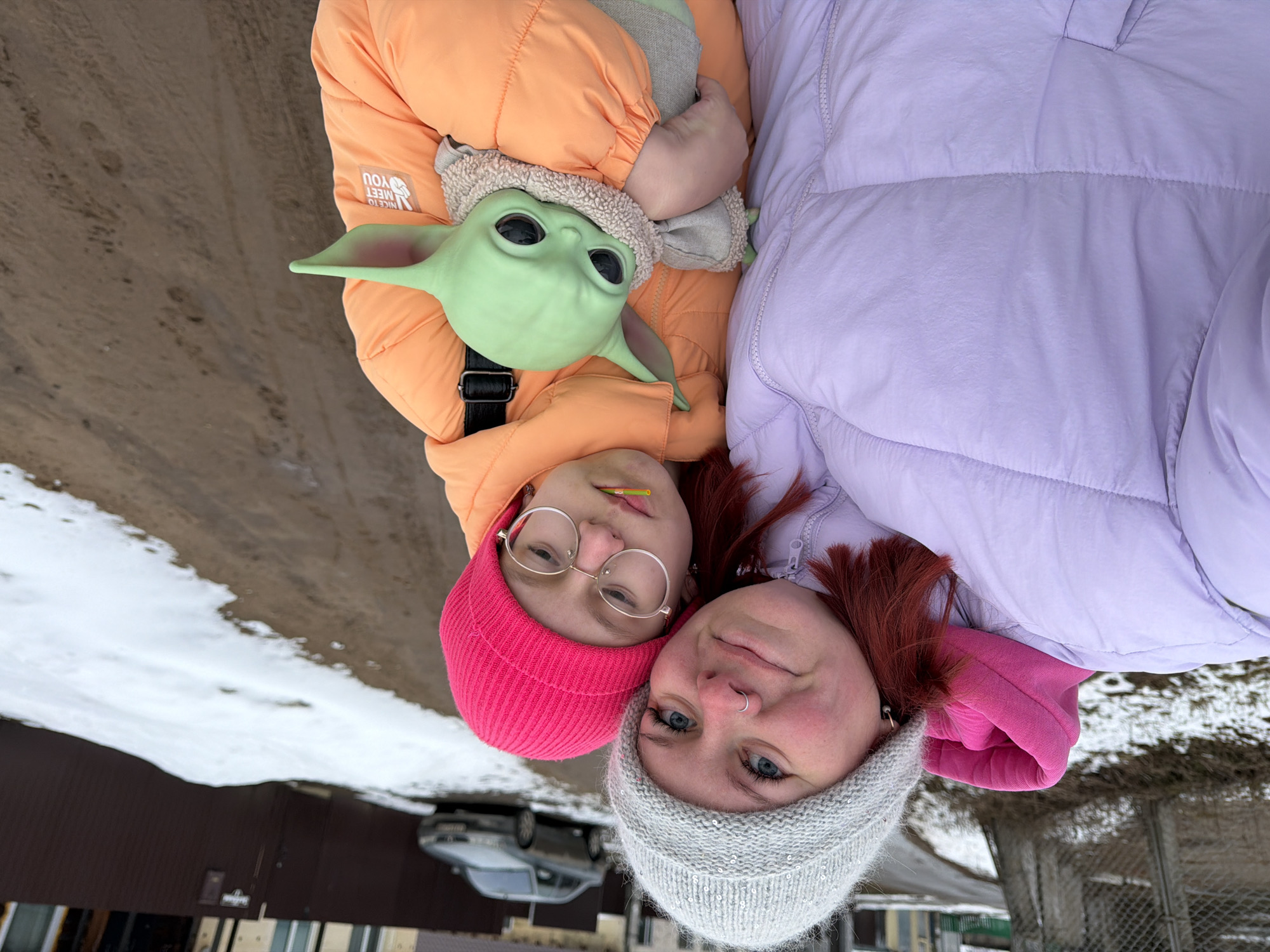 Two women are posing for a selfie outdoors. One woman, wearing a pink beanie, an orange jacket with the phrase "NICE TO MEET YOU" on the sleeve, and glasses, holds a Yoda doll. The other woman, wearing a gray beanie and a purple jacket, is close to the first woman. The background shows a car parked in front of a building and a snow-covered ground near what appears to be a dirt road. Based on the location metadata, the location is identified as the city of Chaikovsky, Russia.