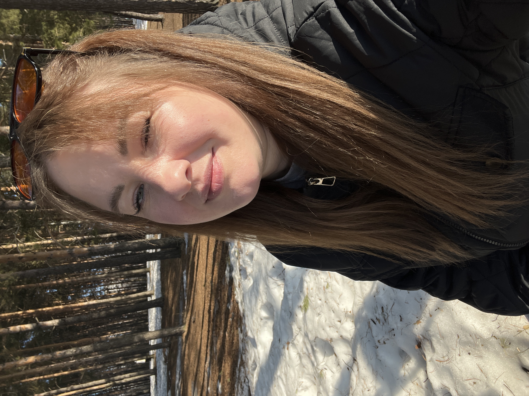 A young adult female, with long, light brown hair and bangs, is depicted in a close-up, selfie-style photograph taken outdoors in Chaikovsky, Russia. She wears amber-tinted sunglasses perched on top of her head. Her left eye is closed in a wink, and a slight smile is present on her face. She is dressed in a dark, possibly black, quilted winter jacket over a grey garment with a visible metallic zipper pull. The background features a dense stand of tall, slender pine trees with reddish-brown trunks. The ground beneath the trees consists of patches of melting white snow interspersed with areas of exposed brown earth and scattered pine needles. The scene is brightly illuminated by direct sunlight, casting distinct shadows across the snow and ground, suggesting a late winter or early spring setting.