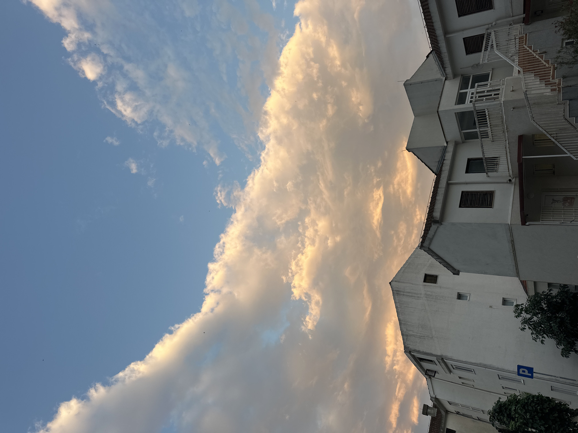 The image shows a street scene in Budva, Montenegro, under a sky filled with clouds illuminated by sunset or sunrise. In the foreground, several white, multi-story buildings are visible. These buildings appear to be residential or institutional, with numerous windows, some with shutters, and balconies. One building has an external staircase with white railings. A blue parking sign with a white 'P' is mounted on a pole to the left of the buildings. Lush green trees partially obscure the lower portions of the buildings. The sky above is a mix of blue and orange hues, with large, fluffy clouds catching the warm light, creating a dramatic backdrop. The overall impression is of a quiet urban environment during twilight.