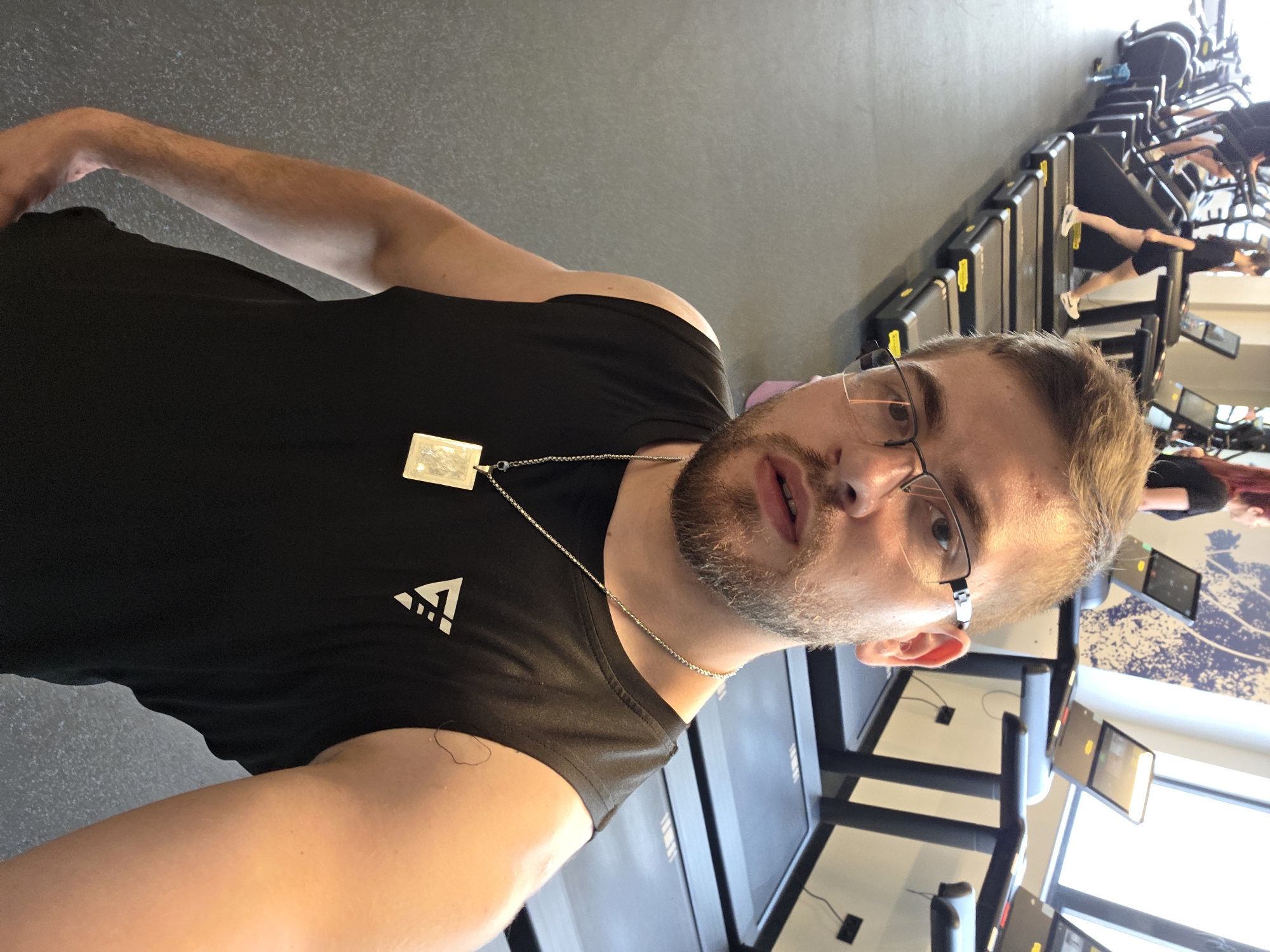 A man with short hair and glasses is taking a selfie in a gym. He is wearing a black t-shirt and a silver necklace. Treadmills are behind him with other people using them. A mural on the wall is visible in the background. The location is not discernible as Plovdiv, Bulgaria based solely on visual evidence.