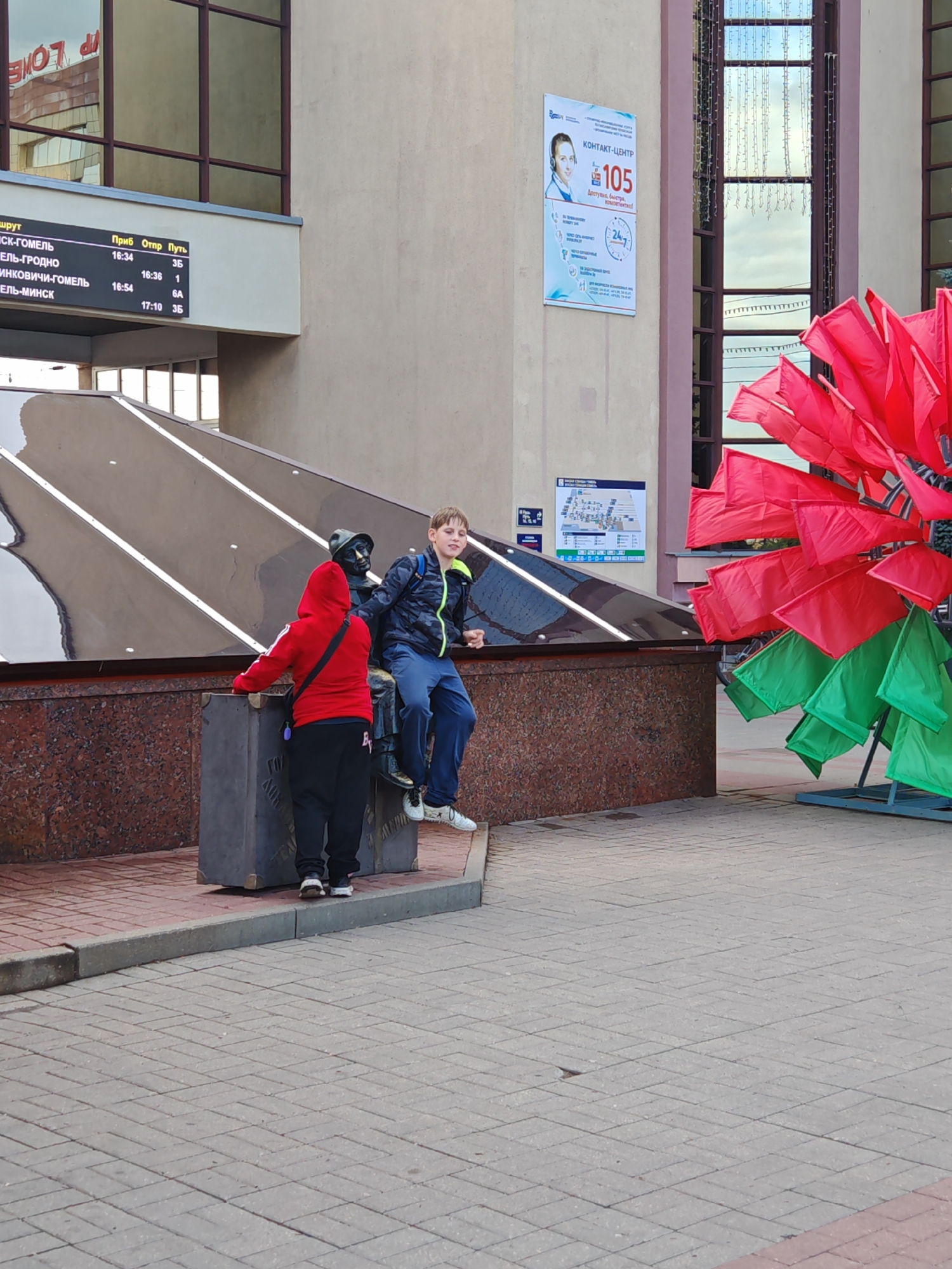 The image depicts an outdoor scene likely in Homyel, Belarus, with a statue of a man wearing a hat and a dark suit, seated on a large suitcase. A young boy wearing a camouflage jacket and dark pants sits beside the statue, looking towards the camera. To the left of the statue, another person, dressed in a red hooded jacket and black pants, stands facing away from the camera, leaning on the suitcase. In the background, a building with large windows and signage is visible. To the right, a large, decorative structure with red and green flags is displayed. A timetable board is also visible on the building's facade. The ground is paved with brick-like tiles.