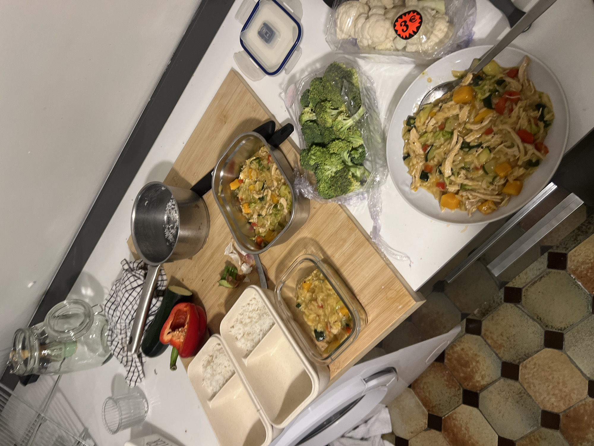 The image shows a countertop scene with food preparation items. A white plate holds a portion of cooked food, possibly chicken and vegetables with a sauce. Packaged broccoli and cauliflower are present, along with containers holding additional portions of the cooked food and rice. A metal pot, cutting board with a knife, a cut red bell pepper, and a zucchini are also visible. The floor tiles suggest the setting is likely in Europe. The "3€" sticker on the cauliflower hints at a location where the Euro is used, such as Paris, France.