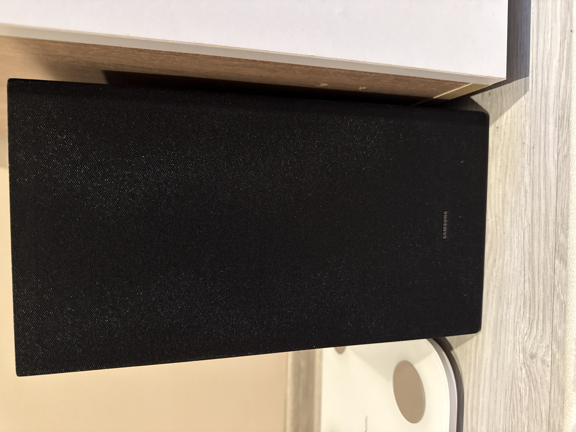 The image shows a black Samsung soundbar resting under a white surface, possibly a shelf. Adjacent to the soundbar is a white scale. The background consists of a light wood-grain surface and a light beige surface. The location is likely indoors. Location in Lovech, Bulgaria cannot be determined from this image.