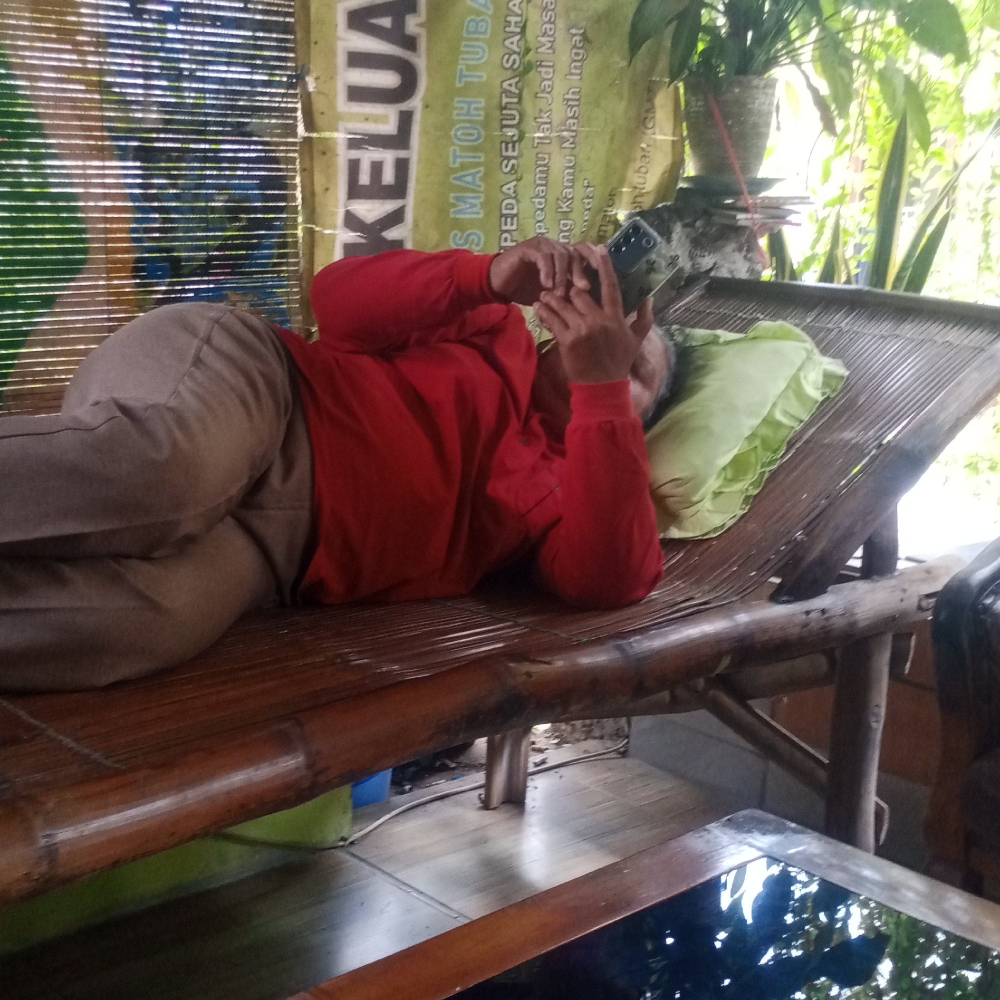 A person in a red long-sleeved shirt and tan pants lies on their left side on a bamboo-slatted daybed. They are holding a smartphone in their right hand, with their head resting on a light green pillow. The background shows a blurred bamboo-slatted screen with a sign that reads "KELUAR". To the right of the daybed, there are potted plants and foliage. The scene appears to be outdoors or in a semi-outdoor setting with natural light filtering through. The floor beneath the daybed is made of polished wood. This location is in Semanding, Indonesia.