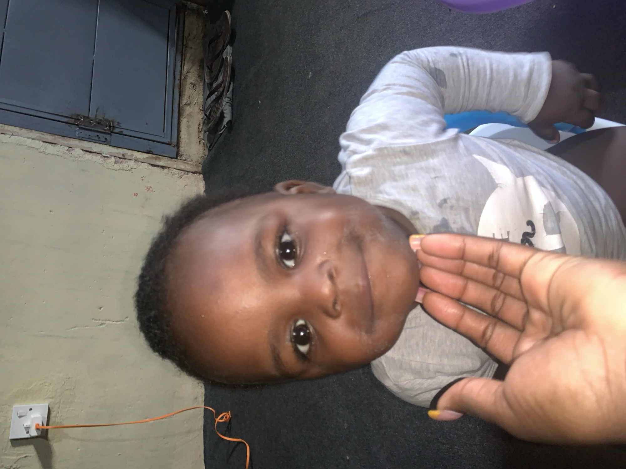 In an indoor setting, a toddler with dark skin and a light grey t-shirt featuring a white graphic of a creature is the central subject. The child's face is turned towards the camera, with dark eyes and a slight smile. A hand, presumably belonging to an adult, is positioned near the toddler's face, with fingernails painted in yellow and pink. The background includes a beige wall with a white electrical outlet and an orange cord, a blue metal door with a lock, and a dark carpeted floor with a pair of dark brown sandals placed near the door. A white plastic object, possibly a chair, is partially visible beneath the toddler. The lighting suggests an indoor environment, possibly a home.