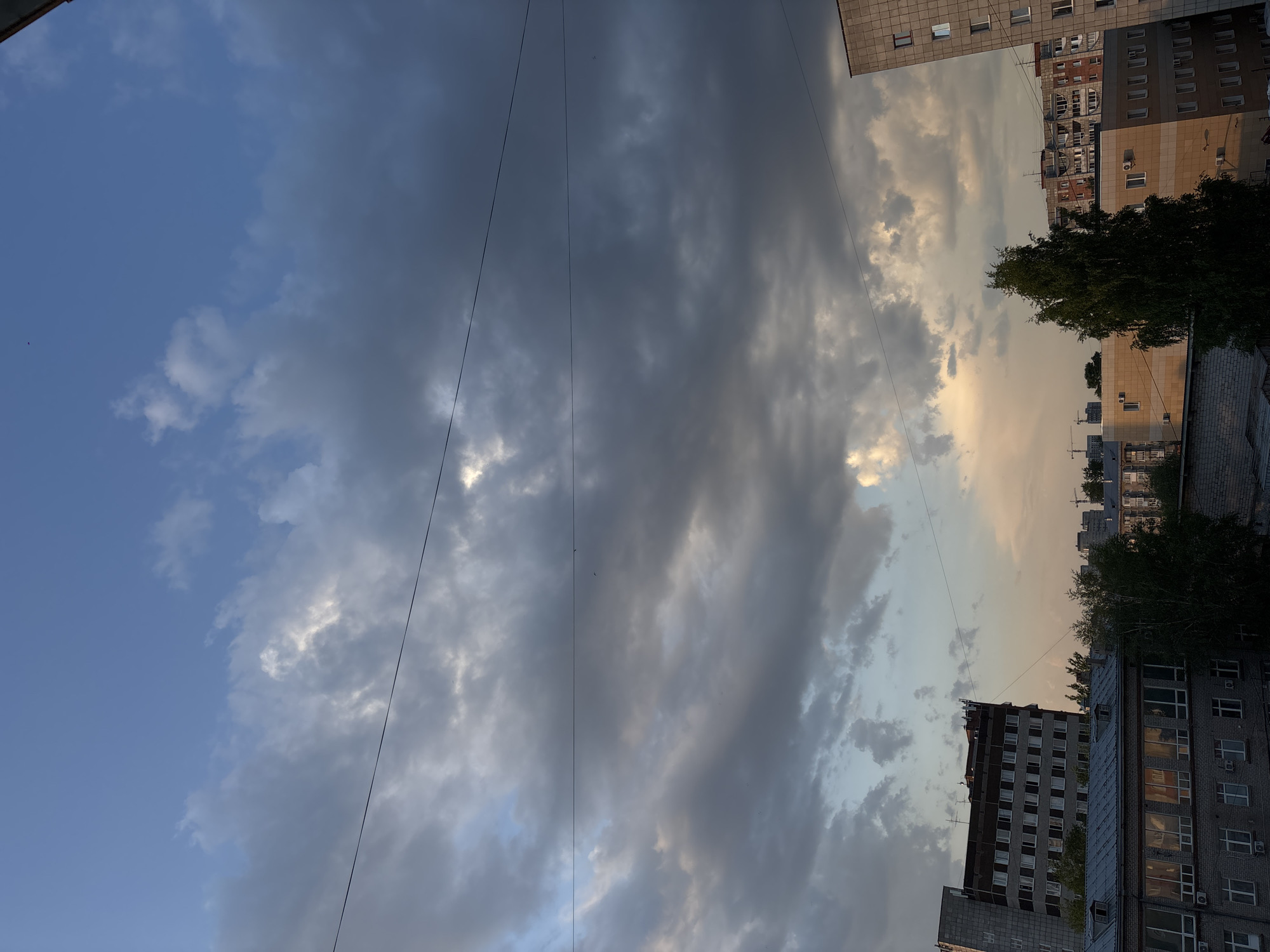 The image depicts an urban scene in Perm, Russia, likely taken in the late afternoon or early evening. The viewpoint is tilted upwards, focusing primarily on the sky and the tops of buildings. The sky is a mix of blue and gray, with patches of sunlight breaking through the cloud cover. Utility lines are visible against the sky. Buildings of varying heights and architectural styles are present, indicating a mixed urban environment. Some buildings are brick with balconies, while others have more modern designs with large windows. Trees are interspersed among the buildings. The overall atmosphere suggests a transition between day and night.