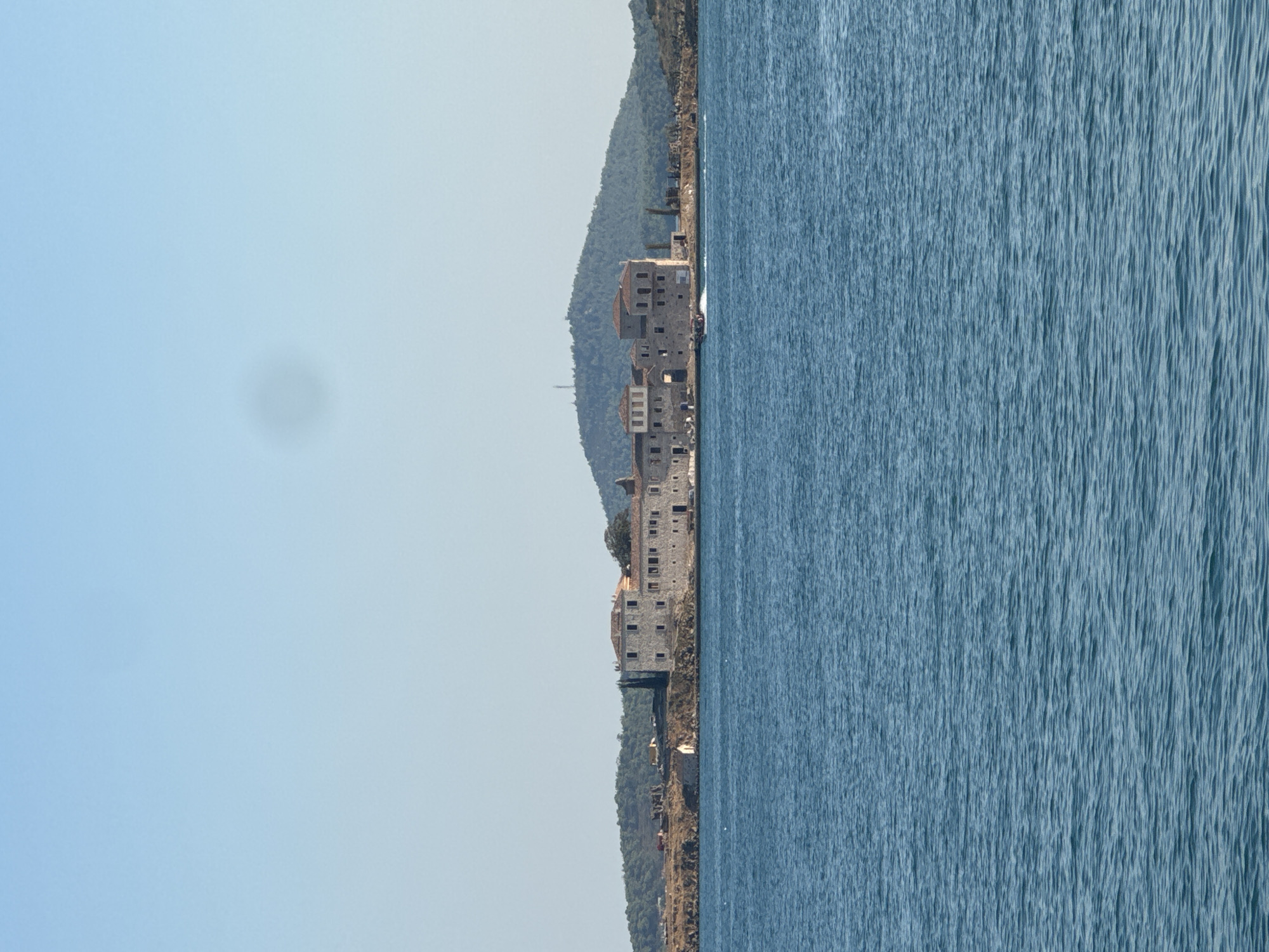 The image depicts a historical stone building complex situated on a rocky island in the foreground, with a body of calm, blue water separating it from the viewer. Behind the island, a lush green, tree-covered hill rises steeply. The building appears to be an old fortress or monastery, characterized by its stone construction, tiled roofs, and multiple windows. A small, fast-moving boat with several people on board is visible in the water, leaving a white wake. The sky is a clear, pale blue, with a faint circular blur in the upper portion of the frame, possibly a lens artifact. The overall impression is one of a serene, historical coastal landscape. This building complex is located on the island of Cunda (also known as Alibey Island) in Ayvalık, Turkey.