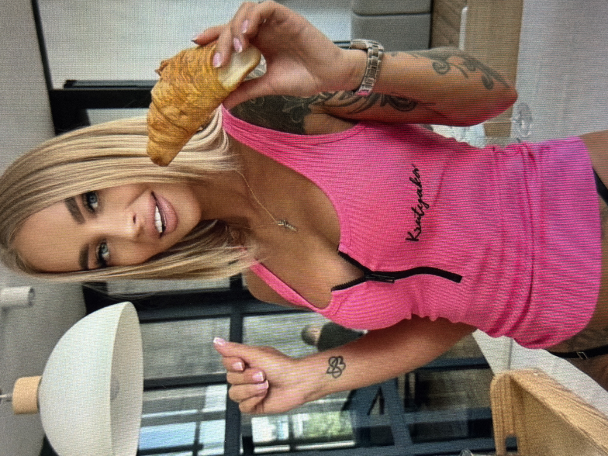 In a brightly lit indoor setting, a woman with blonde hair and blue eyes is captured in a close-up shot, holding a croissant. She is wearing a vibrant pink, ribbed tank top with a zipper detail and has visible tattoos on her arms and a small tattoo on her left wrist. The background includes a modern light fixture on the ceiling and a blurry glimpse of what appears to be an interior space. There is no information in the image to indicate a location in Shumen, Bulgaria.
