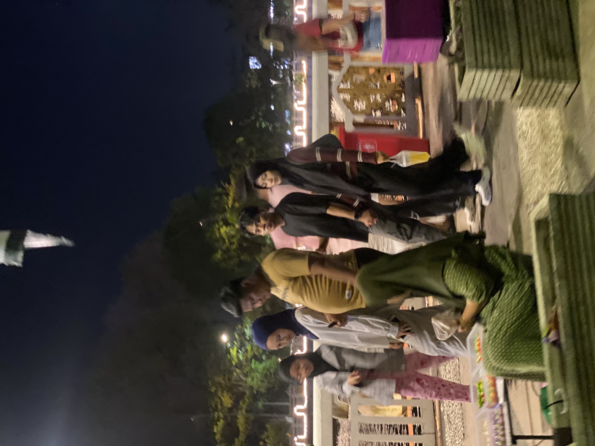 The image captures a nighttime scene in Banjarmasin, Indonesia, likely along a waterfront promenade or bustling street. Several individuals are walking, suggesting a public gathering or a casual outing. One person, a woman in a green hijab and dress, is seated and appears to be selling sweets from clear plastic containers arranged on a table. In the background, decorative lights adorn a railing, illuminating the pathway and creating a festive atmosphere. The presence of trees and the dark sky indicate an outdoor setting at night.
