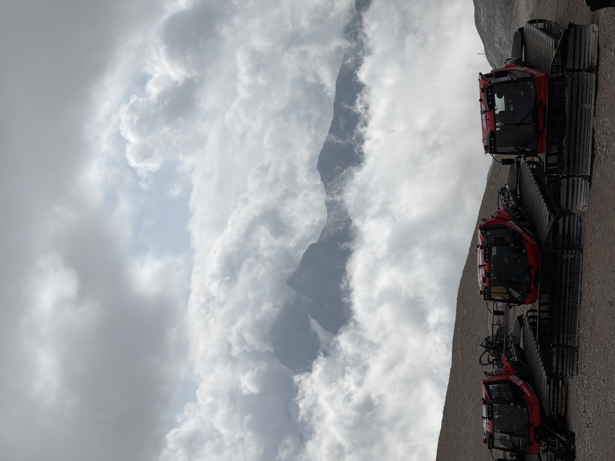 Three red snow groomers are parked on a gravelly slope with a dramatic cloudy sky and mountain peaks visible through the clouds. The snow groomers are large, tracked vehicles with enclosed cabins. They are positioned in a line from left to right, with the leftmost groomer partially obscured by the other two. The sky is filled with heavy, gray clouds, with patches of lighter sky visible. The mountain peaks are dark and rugged, appearing to be shrouded in mist or clouds. The foreground is a mix of small rocks and gravel. The overall impression is a dramatic and powerful landscape. The location is Zugspitze, the highest peak in Germany, which is accessible from Garmisch-Partenkirchen.