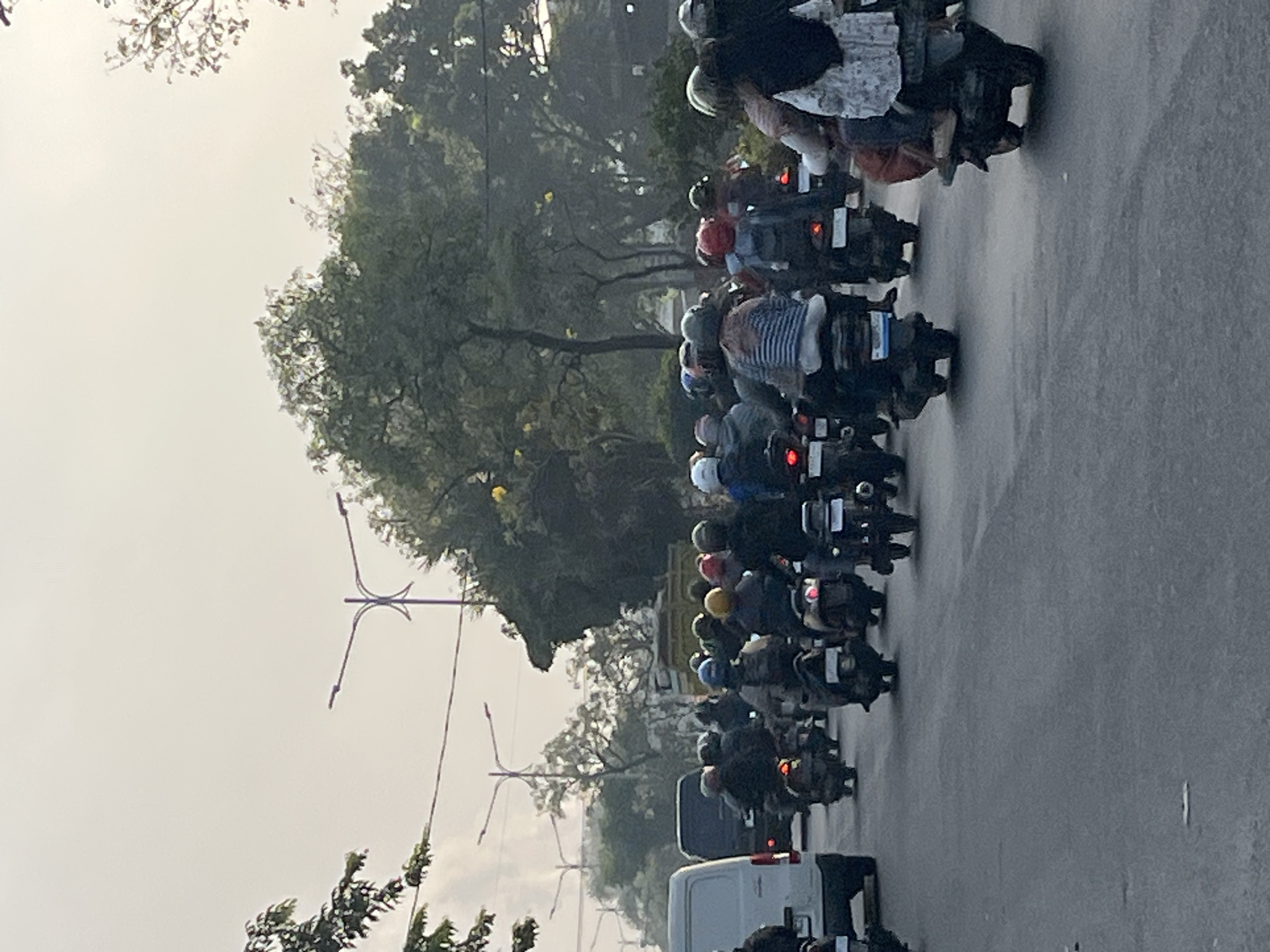 The image depicts a congested street scene in Gelam, Indonesia, with a large group of motorcyclists riding in the same direction. The motorcyclists are diverse, with many wearing helmets and some carrying passengers. The vehicles are primarily scooters and motorcycles, with a white van visible on the left side of the frame, possibly indicating a delivery or commercial vehicle. The environment appears to be a typical urban setting with trees lining the street and utility poles in the background. The sky is overcast, suggesting a cloudy or hazy day. The overall impression is one of busy, everyday traffic in a Southeast Asian city.