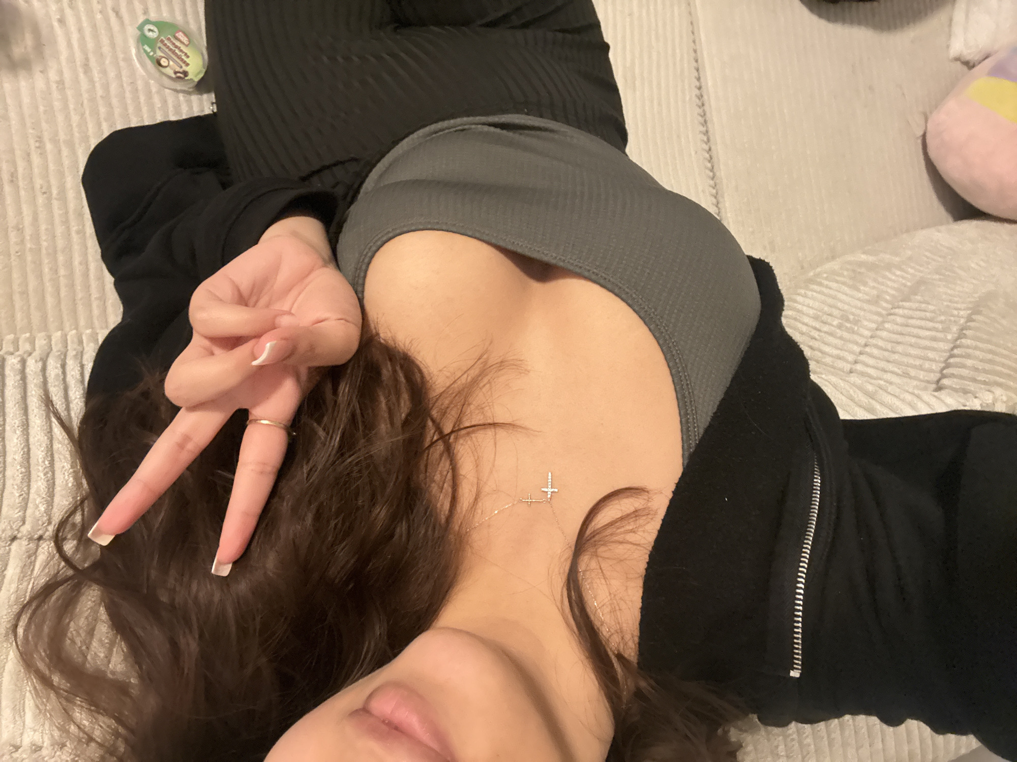 A person is lying on a gray textured surface, possibly a bed or couch. They are wearing a gray top, a black zip-up hoodie, and dark ribbed pants. The person's right hand is raised, forming a "peace" sign. They have long brown hair and are wearing a gold ring on their index finger. A necklace with cross pendants is visible. A small, circular container with a green label is located in the upper left corner. A pink and yellow stuffed animal is visible in the upper right corner. There is no information available about the location in Mönchengladbach, Germany.