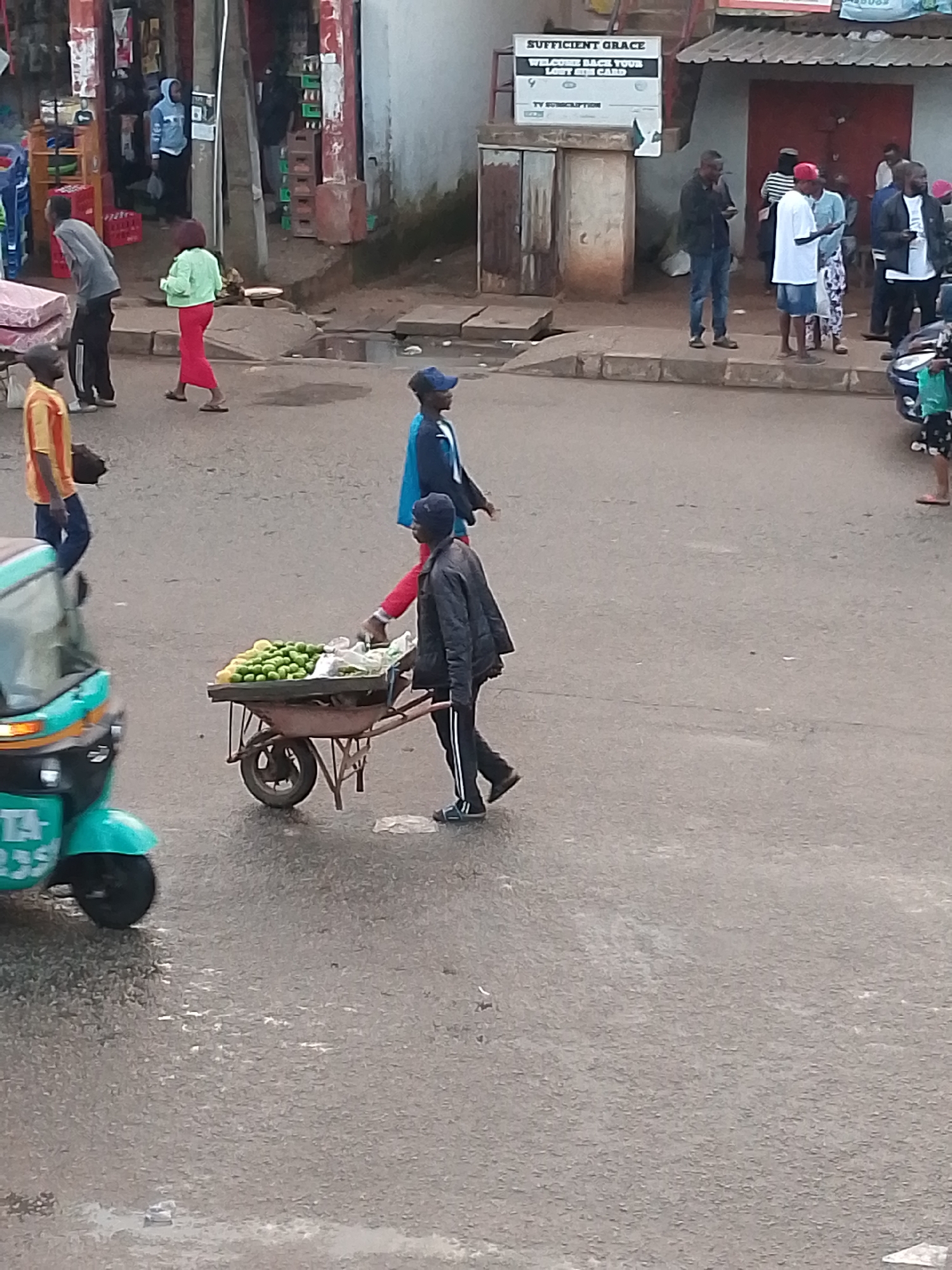 The image depicts a street scene in Bukuru, Nigeria. In the foreground, a man pushes a wheelbarrow laden with produce, likely limes or lemons, and some plastic bags. He is wearing a dark jacket and black pants with white stripes. Behind him, to the left, a teal-colored auto-rickshaw is partially visible. Further back, various people are standing and walking along the sidewalk. To the far left, a person in a grey shirt and black pants walks away from the camera. A woman in a green top and red pants is walking towards the right. To the right of the wheelbarrow, a person in a blue jacket and blue cap walks by. In the background, there are buildings, including a shop with a sign that reads "SUFFICIENT GRACE WELCOME BACK YOUR LOST SIM CARD". Several individuals are gathered on the sidewalk in front of the shops. The ground appears to be paved asphalt and wet in places, suggesting recent rain.
