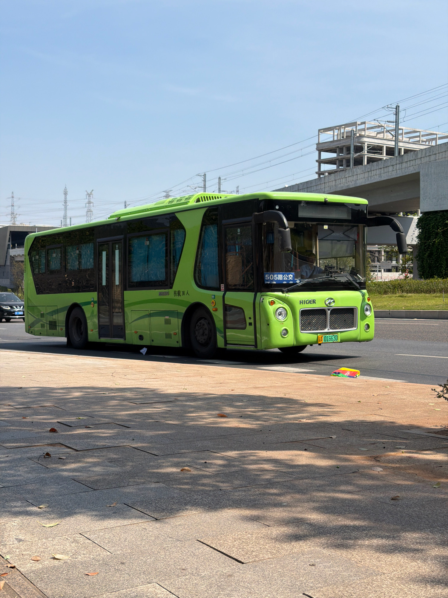 A bright green Higer bus is parked on the side of a road in Jiangjin, China, with its front facing the viewer's right. The bus has "505路公交" and "载38人" written on its front, indicating it is bus route 505 and can carry 38 passengers. The license plate number is "A 01663D". To the left of the bus, a dark-colored sedan is visible. The bus is positioned on an asphalt road adjacent to a paved walkway, where several colorful, flat objects resembling toys or promotional items are scattered. The environment features overhead power lines and a concrete overpass structure in the background. The sky is clear and blue, and the scene is illuminated by bright sunlight, casting shadows on the walkway.