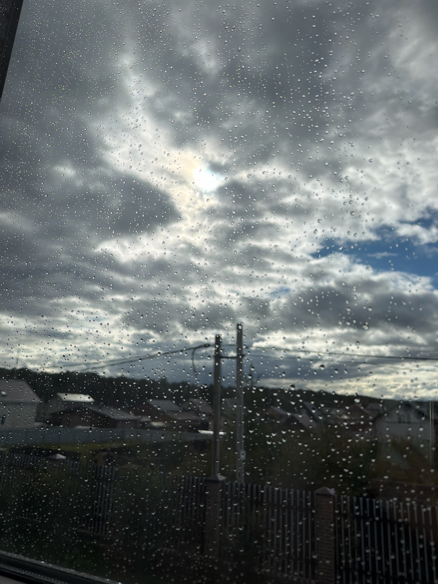 The image depicts a view through a rain-streaked window overlooking a suburban landscape. The sky is overcast with a mix of dark and lighter gray clouds, with a patch of sunlight breaking through in the upper central area. Below the sky, a dark, silhouetted panorama of houses with tiled roofs is visible, interspersed with trees and vegetation. A dark, vertical fence runs across the mid-ground, obscuring some of the view. In the foreground, numerous raindrops cling to the glass, distorting the scene and creating a textured effect. There are no people or animals visible in the image. The scene appears to be set in a residential area.