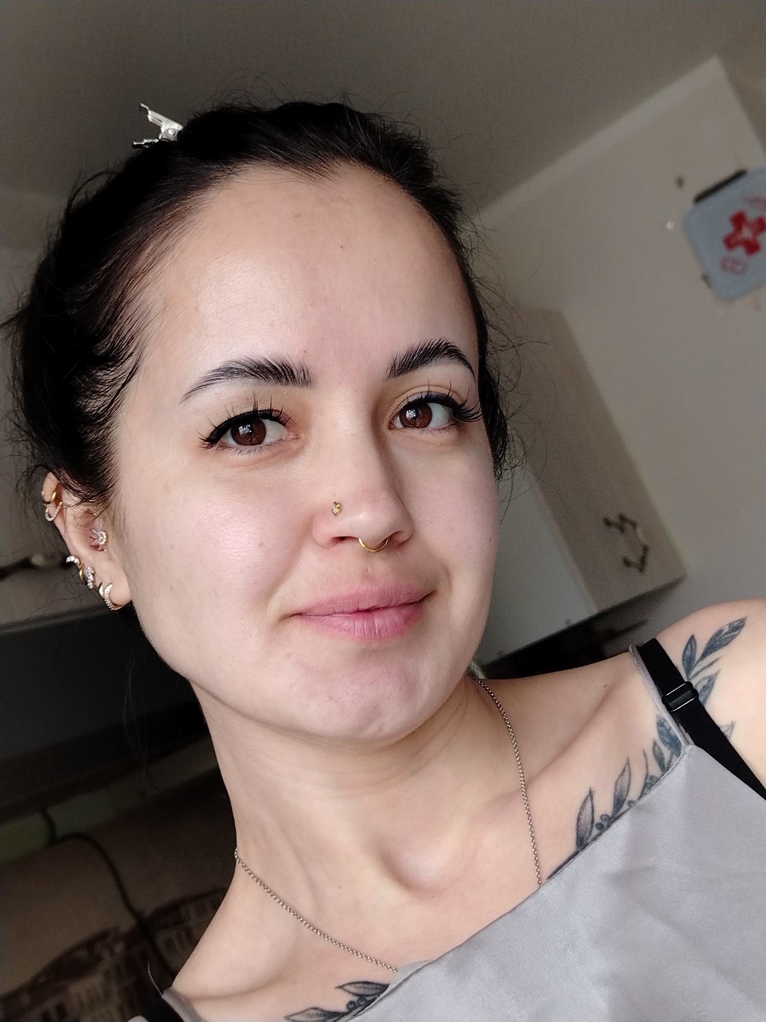 A selfie of a young woman indoors. She has dark hair pulled back, brown eyes, and light skin. She is wearing a nose ring and multiple earrings on one ear. A thin silver necklace is around her neck. She has a leafy tattoo on her shoulder and another one on her chest. The background is blurry, showing a white cabinet and a first aid kit hanging on the wall. There is no indication in the picture that the image was taken in Noviy, Russia.