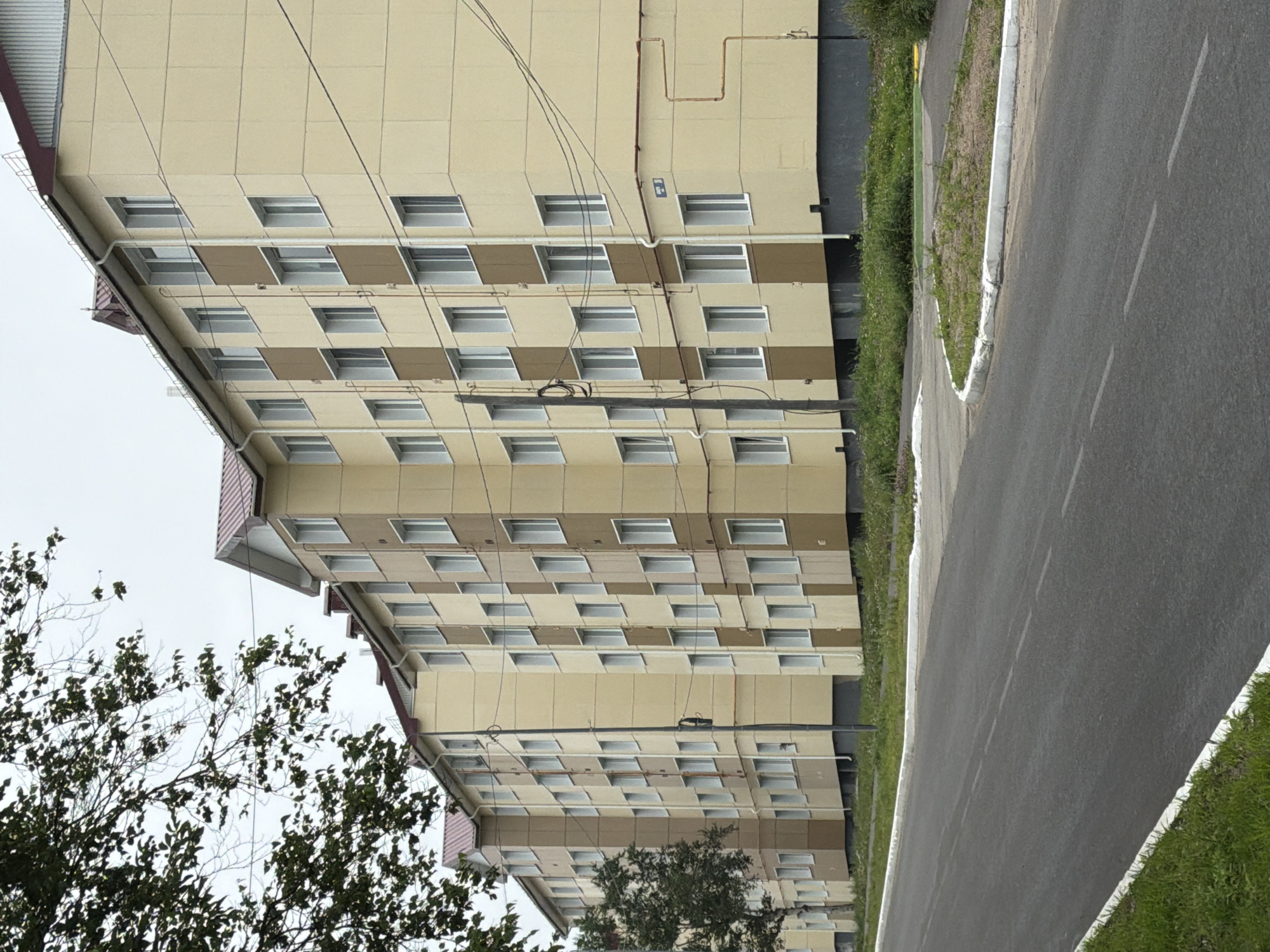 A low-angle, wide shot captures a segment of a long, multi-story apartment building, painted in alternating bands of pale yellow and light brown. The building features a dark grey foundation and a reddish-brown metal roof with white gutters and downspouts. Numerous windows with white frames punctuate the facade. The building is bordered by a narrow strip of green grass and wildflowers at its base. A paved road with white lane markings runs diagonally across the foreground, with a white concrete curb and a grassy shoulder along its edge. A mature tree with dark green foliage extends into the upper left corner of the frame. Overhead, a network of utility wires is visible against an overcast sky. A tall, dark pole with electrical equipment stands in front of the building. The scene depicts a residential area in Okha, Russia, under a cloudy sky.