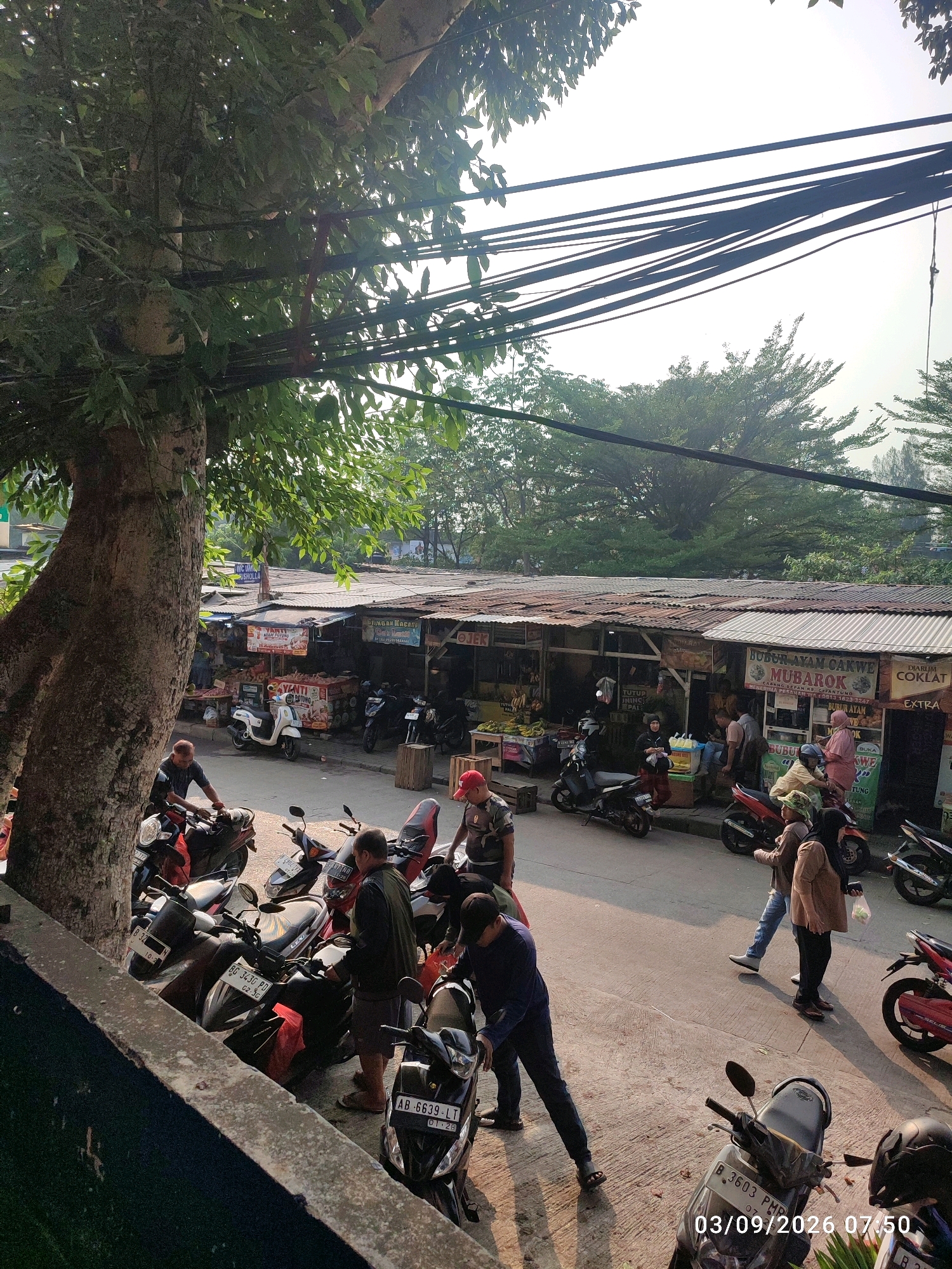 This image captures a bustling street scene in East Jakarta, Indonesia, likely a market area with several vendors. The foreground is dominated by a large tree on the left, with its branches and leaves partially obscuring a collection of parked motorcycles. In the middle ground, a row of small stalls with corrugated metal roofs line the street, displaying various goods, possibly food and produce. People are engaged in various activities: some are walking, some appear to be interacting with vendors, and others are near their motorcycles. Several motorcycles are parked along the street, indicating a common mode of transportation. Power lines crisscross the upper portion of the image, casting shadows on the sky. The overall impression is one of a lively, everyday commercial environment.