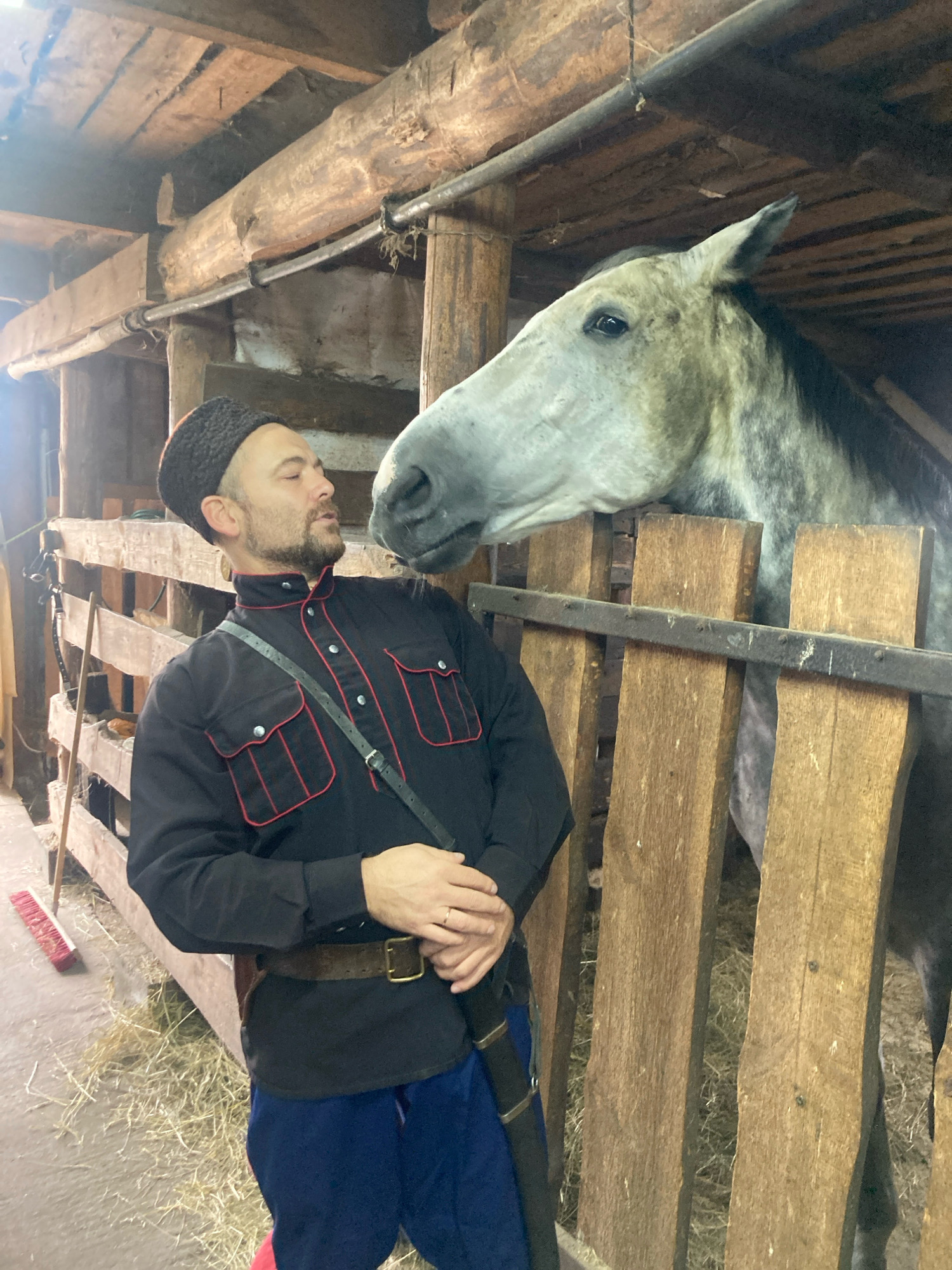 In a rustic barn with wooden beams and hay on the floor, a man dressed in a black traditional Cossack-style outfit with red trim and a fur hat stands next to a light gray horse. The man is wearing a black shirt with two chest pockets, a dark belt with a brass buckle, and blue pants with a red stripe. He has a beard and is looking at the horse, which is partially visible over a wooden fence. The horse's head is lowered, appearing to be close to the man. The environment suggests an agricultural or historical reenactment setting within the Тэльмінскі сельскі Савет, Belarus.