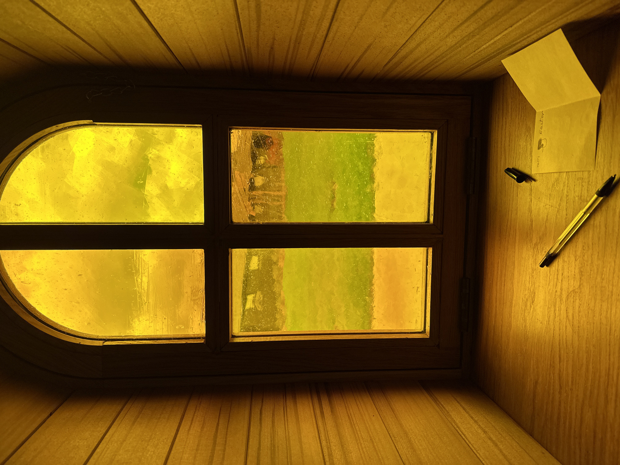The image displays the interior of a small, wooden-paneled structure located in Maldon, United Kingdom. The walls are constructed from horizontal light-colored wooden slats, illuminated by a warm yellow light. A central, wooden-framed window, consisting of an arched upper section and a rectangular lower section, is divided into four panes by a horizontal bar and a vertical mullion. All glass panes are textured and appear tinted yellow or amber, exhibiting numerous water droplets on their exterior surfaces. Through the lower panes, a green grassy area is visible, with indistinct darker forms, possibly structures, in the background. The upper panes show a more abstract yellow view, consistent with filtered sky or distant elements. Directly beneath the window, on a flat wooden sill, a transparent yellow ballpoint pen with a black end cap rests horizontally. Adjacent to it on the right, a separate black pen cap is present. Further to the right, a small, folded yellow card lies open, displaying handwritten text in dark ink that reads "Happy Easter" accompanied by a small, simple drawing resembling a cap.