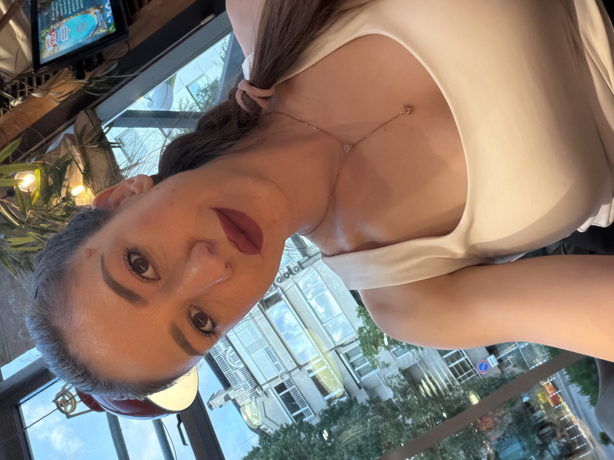 The image is a close-up selfie of a woman with medium skin tone, dark hair pulled into a ponytail secured with a light pink scrunchie. She has prominent eyebrows, eyeliner, and dark red lipstick. A small raised mark or blemish is visible above her right eyebrow. She is wearing a white or off-white sleeveless top and a rose gold-toned lariat necklace featuring a small triangular pendant near the collarbone and another small, possibly heart-shaped or geometric pendant dangling lower.
The scene appears to be an indoor setting, likely a cafe or restaurant, characterized by warm ambient lighting from various fixtures, including a red, industrial-style dome lamp hanging from the ceiling. Greenery and wooden structural elements are also visible. In the upper right corner, a small screen displays what appears to be a graphic with "HAPPY BEACH" text.
Through a large glass window behind the woman, an urban streetscape is visible. Multi-story buildings with balconies, characteristic of residential or commercial architecture in Sofia, Bulgaria, line the street. One building features prominent white decorative railings. Trees with green foliage are present along the street. A blue circular road sign with a single red diagonal line, indicating "No Parking" in European road signage, is clearly visible. The sky is light blue, suggesting daytime or early evening. The overall environment is consistent with an urban area in Sofia, Bulgaria.