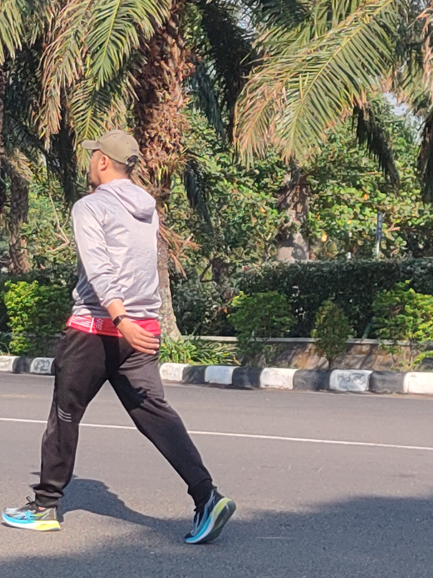 A man wearing a grey hooded jacket, black sweatpants, a green baseball cap, and running shoes is walking on a paved road. He has a red belt around his waist. He is outdoors, with palm trees and lush green foliage in the background. The scene appears to be in Ngasem, Indonesia. The man is walking from left to right, with his left leg forward. His shadow is cast on the road behind him. The road has a white line marking and a curb with black and white blocks.