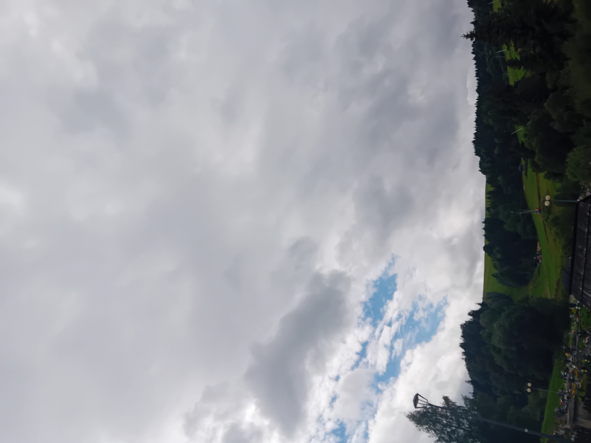 This image depicts a cloudy day in Zuberec, Slovakia. The foreground shows a cemetery with gravestones, a lamp post, and trees. In the background, a green, grassy slope is visible, dotted with trees and a few small structures. The top of the image is dominated by a gray, overcast sky with scattered patches of blue sky and lighter clouds. The scene appears to be a somewhat somber or reflective atmosphere due to the cemetery setting.