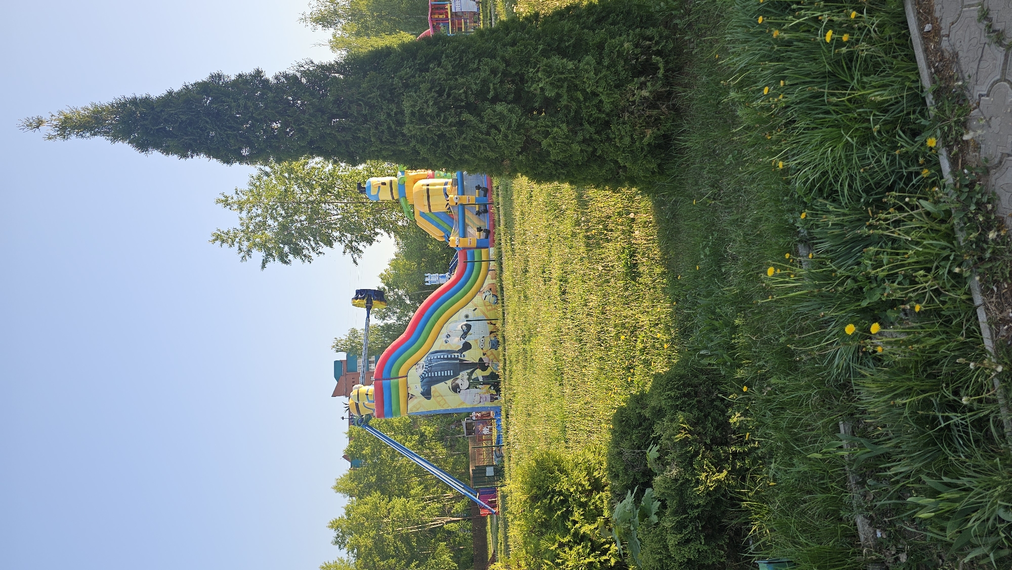 The image shows a park scene with a large inflatable slide featuring a rainbow design and images of cartoon characters. The slide is positioned on a green lawn, adjacent to a tall, narrow evergreen tree on the left and a patch of green grass and dandelions on the right. In the background, there are other trees and buildings. The sky is clear and blue. The location is in an unknown city, in an unknown region.
