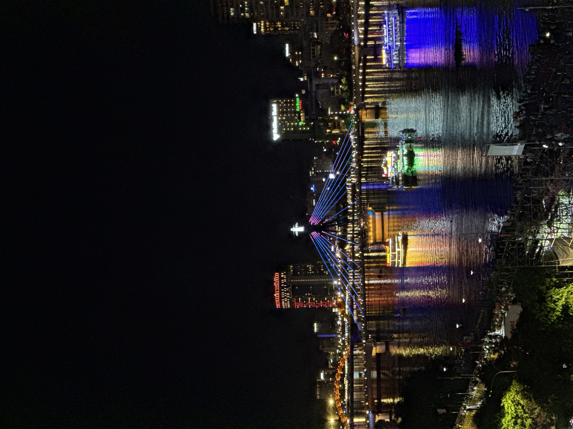 The image captures a nighttime view of the Han River in Da Nang, Vietnam, featuring the Dragon Bridge and surrounding cityscape. The Dragon Bridge, illuminated with a vibrant blue light and adorned with multi-colored lights along its cables, dominates the central portion of the frame. Atop the bridge, a statue, possibly of Jesus Christ, is visible, illuminated by a white light.
Several boats are present on the river. One is a brightly lit yellow boat with a decorative top, reflecting yellow and orange light onto the water. Another boat, colored green and white, is also illuminated and reflects light onto the river. A larger boat, decorated with purple lights, is seen on the right side of the frame, its purple reflection stretching across the water.
The riverbanks are lined with buildings, many of which are illuminated with various colors. A tall, prominent building with red lights along its upper levels stands out. Another building displays a green illuminated sign with the text "Brilliant Hotel" and "ANDOVER HOTEL" in red below it. Reflections of the city lights and the boats are visible on the rippling surface of the Han River, creating a colorful and dynamic display.
Along the lower left and bottom edges of the image, a dense crowd of people is gathered on the riverbank, suggesting a public event or gathering. Some are silhouetted against the lights, while others are partially visible. Trees with green foliage are also present in the foreground on the left. The overall scene depicts a lively urban waterfront at night, characterized by striking bridge illumination, vibrant reflections, and a visible human presence.