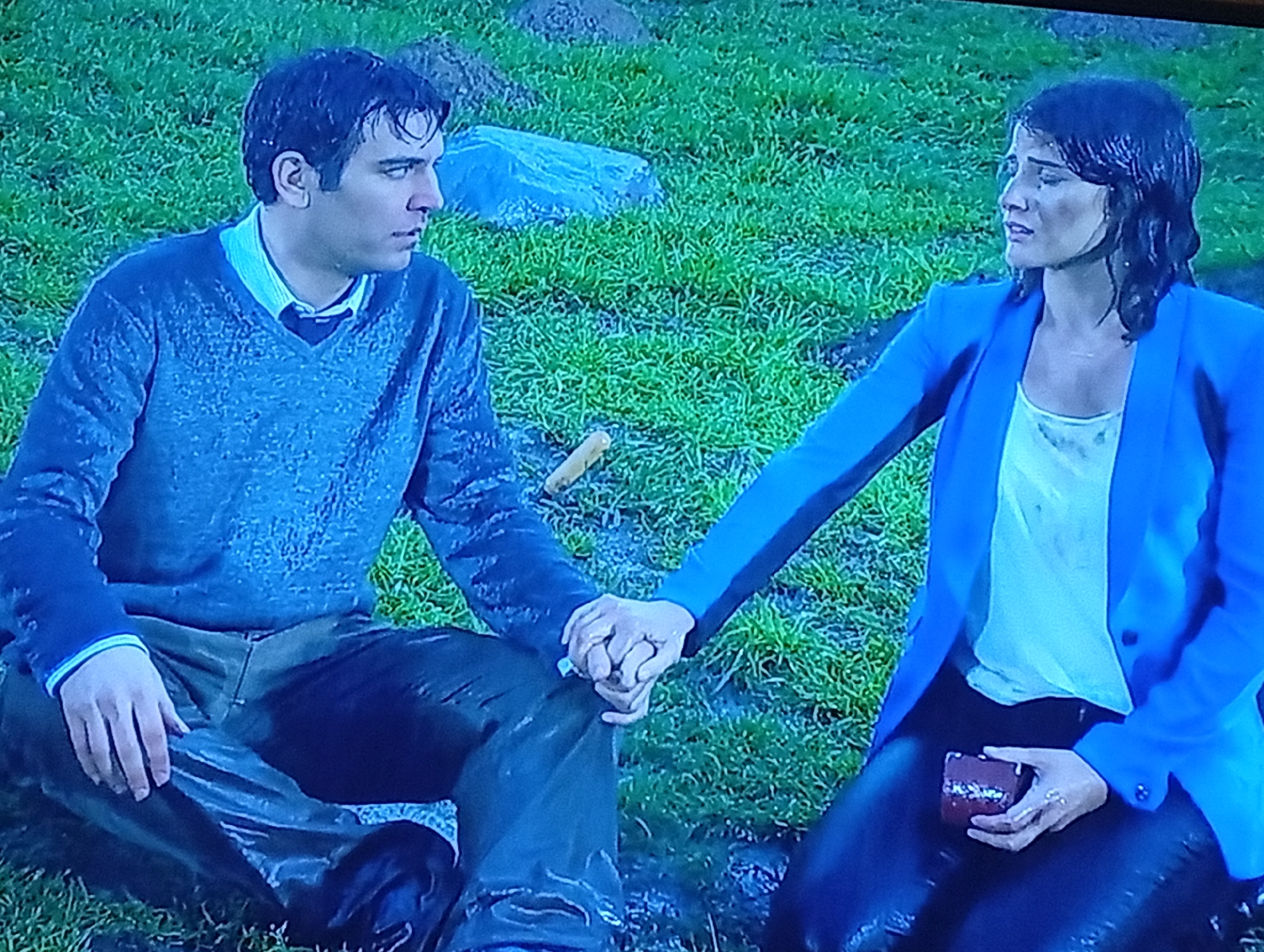 A man and a woman are sitting close together in a grassy area with visible moisture, possibly from rain. The man is seated in a cross-legged position, wearing a v-neck sweater over a collared shirt and darker-colored pants. The woman, also sitting on the ground, is wearing a white shirt under a blue blazer with dark pants. They are holding hands. The woman is holding a small, dark red box. The background is mostly green grass, with a few rocks and soil patches visible. The location appears to be a park or other open space. The city and other locations are unknown.