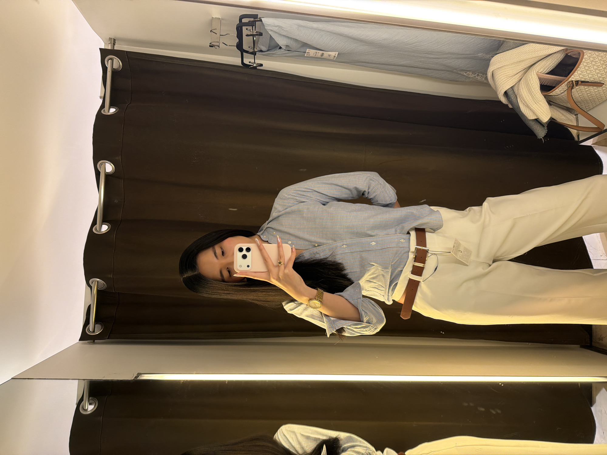A person is taking a selfie in a fitting room. The fitting room has a mirror, a brown curtain, and clothing racks with clothes.