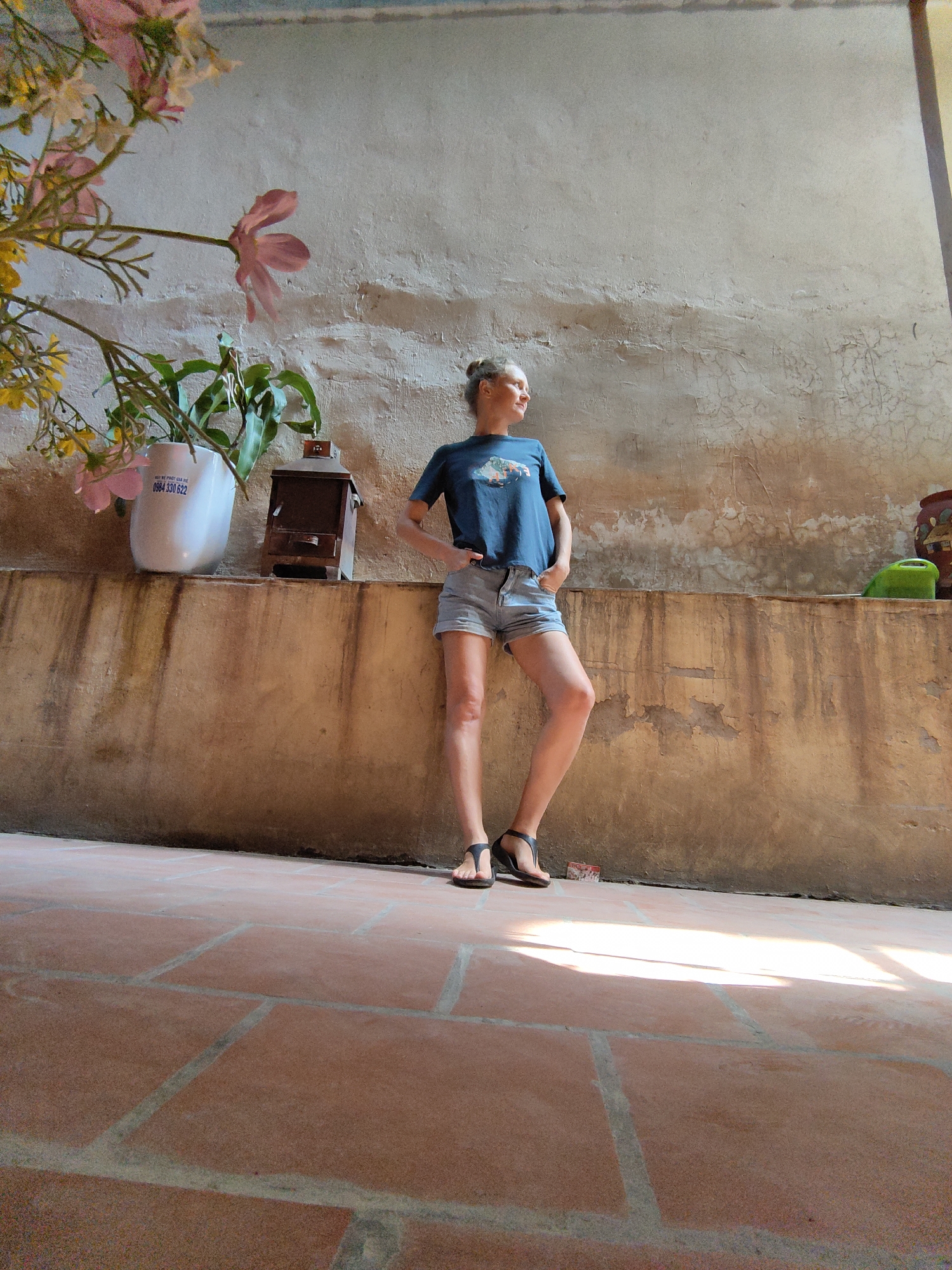 A woman stands in a courtyard in Hà Nội, Vietnam. She wears denim shorts, a t-shirt, and sandals. She leans against a stucco wall, with her hands in her pockets. Various objects sit atop the wall, including a white flower pot, an antique box, and a green watering can. The courtyard floor is made of terra cotta tiles. A flowering plant is in the foreground.