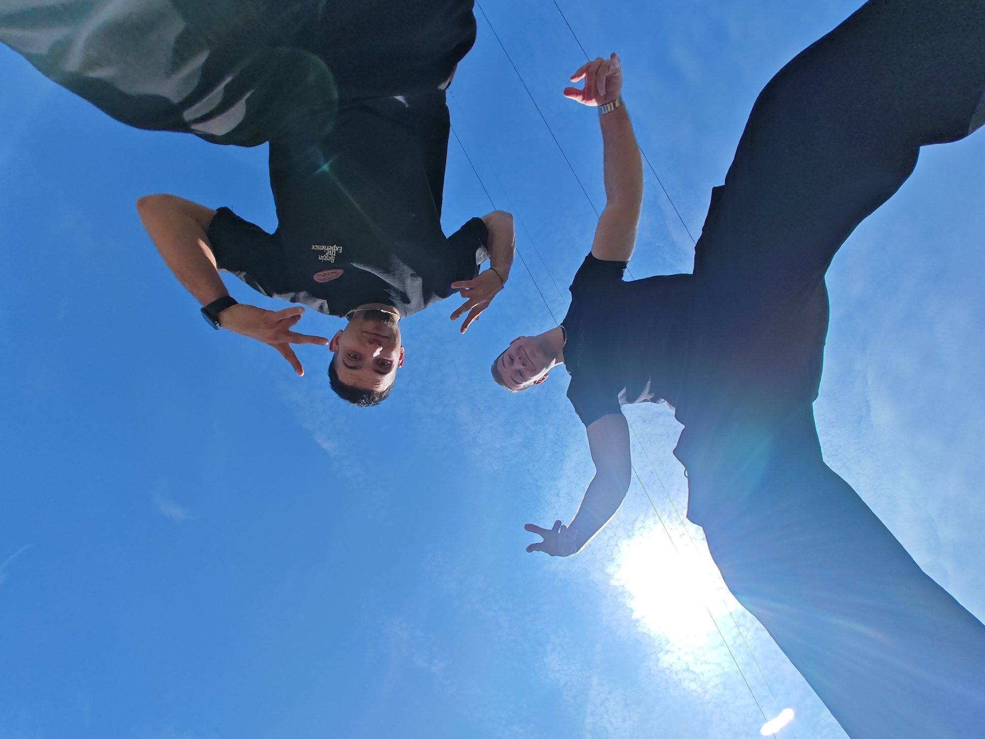 Two individuals are depicted from a low-angle perspective against a bright blue sky with scattered wispy clouds, likely in an outdoor area of Shumen, Bulgaria. The specific location within the city cannot be determined from the visual information provided. The sun is visible in the lower right, casting lens flares.
Both subjects appear to be adult males, positioned towards the top of the frame, looking down towards the camera. They are dressed in black short-sleeved t-shirts.
The individual on the left has dark hair and a light complexion. He wears a dark wristwatch on his left wrist and a light-colored bracelet on his right wrist. His t-shirt features a circular logo with the text "Begin The Experience". He makes a "peace sign" gesture with his left hand, and his right hand is partially curled with fingers extended downwards. He exhibits a slight smile.
The individual on the right has lighter hair and a light complexion. He wears a metallic-colored watch or bracelet on his left wrist. His left arm is raised with a partially curled hand, fingers extended downwards, and his right arm is bent with a similar hand gesture. He also displays a slight smile.
Faint thin lines, possibly power lines or overhead wires, are visible crossing the sky between the two subjects.