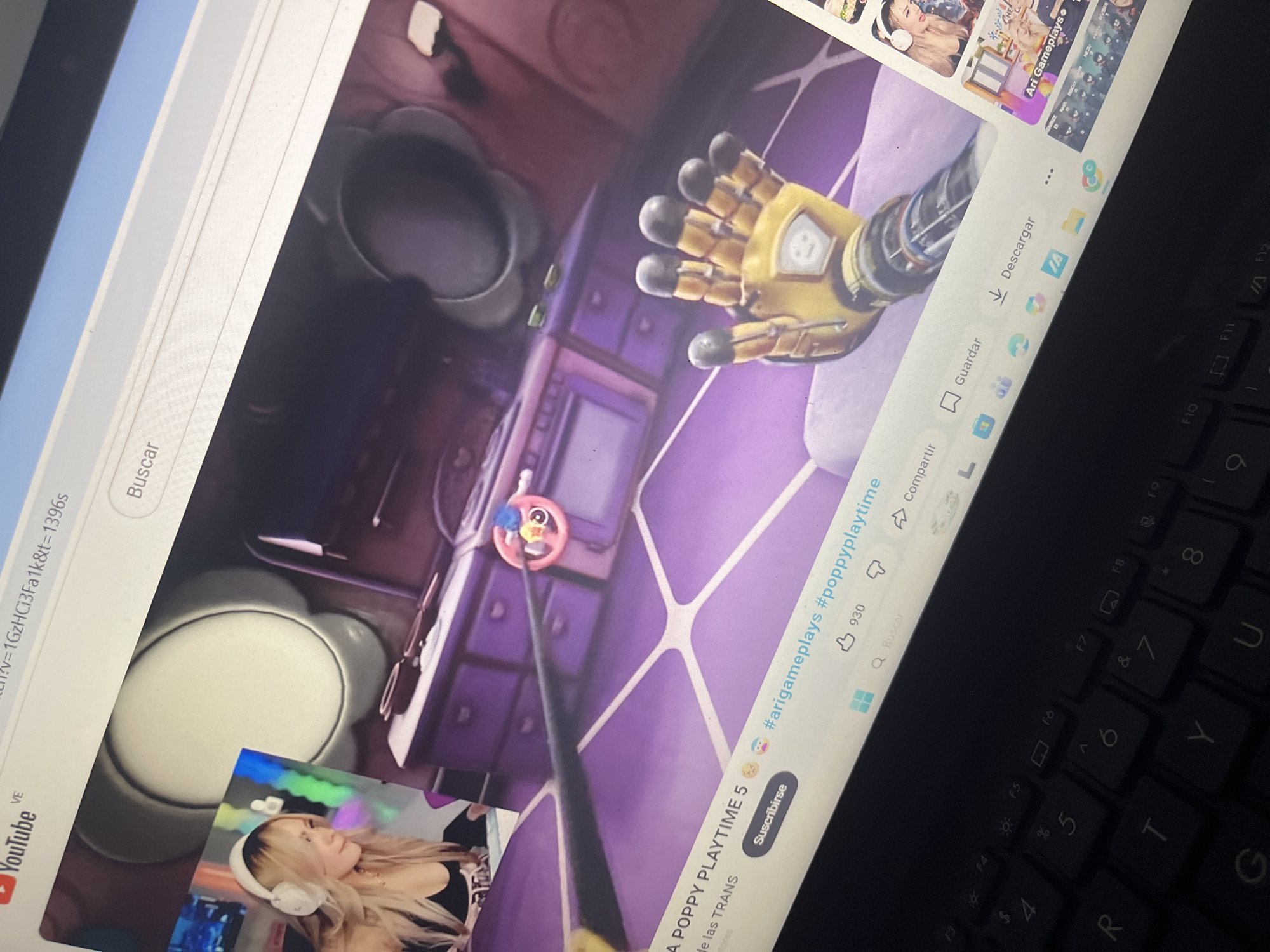The scene is depicted as occurring in Santa Ana, Venezuela. The primary subject is a dark-colored laptop, presented from an elevated, slightly angled perspective, with its screen displaying a YouTube video.
The laptop screen shows a video game, identified by hashtags as "#arigameplays #poppyplaytime". The game interface features a first-person perspective within a purple-hued room. In the foreground, a prominent yellow robotic hand with black fingertips and a smiley-face emblem on its wrist is visible, extending towards the viewer. In the mid-ground, a small, pink, circular object with blue and yellow elements, resembling a toy, is present. The background shows purple cabinets or drawers with small handles.
A smaller picture-in-picture window in the upper left corner of the screen displays a female streamer with long blonde hair, wearing a dark top and white over-ear headphones. She is looking towards the left side of the frame.
The YouTube interface includes the "YouTube" logo and "VE" in the top left. The URL in the address bar is partially visible as `v=1GzHCI3Fa1k&t=1396s`. A search bar labeled "Buscar" (Spanish for search) is located above the video. Below the video, a "Suscribirse" (Subscribe) button is visible alongside the text "A POPPY PLAYTIME 5" and the hashtags. The video has 930 likes. Below the video, icons for sharing ("Compartir"), saving ("Guardar"), and downloading ("Descargar") are visible. A sidebar of recommended videos on the right includes a thumbnail with "Ari Gameplays" visible.
The laptop's black QWERTY keyboard is partially visible in the lower part of the image, appearing dimly lit, with function keys (F3 to F9) and standard alphanumeric keys. The surrounding environment is dark, suggesting the image was captured in a low-light setting where the laptop screen is a primary light source.