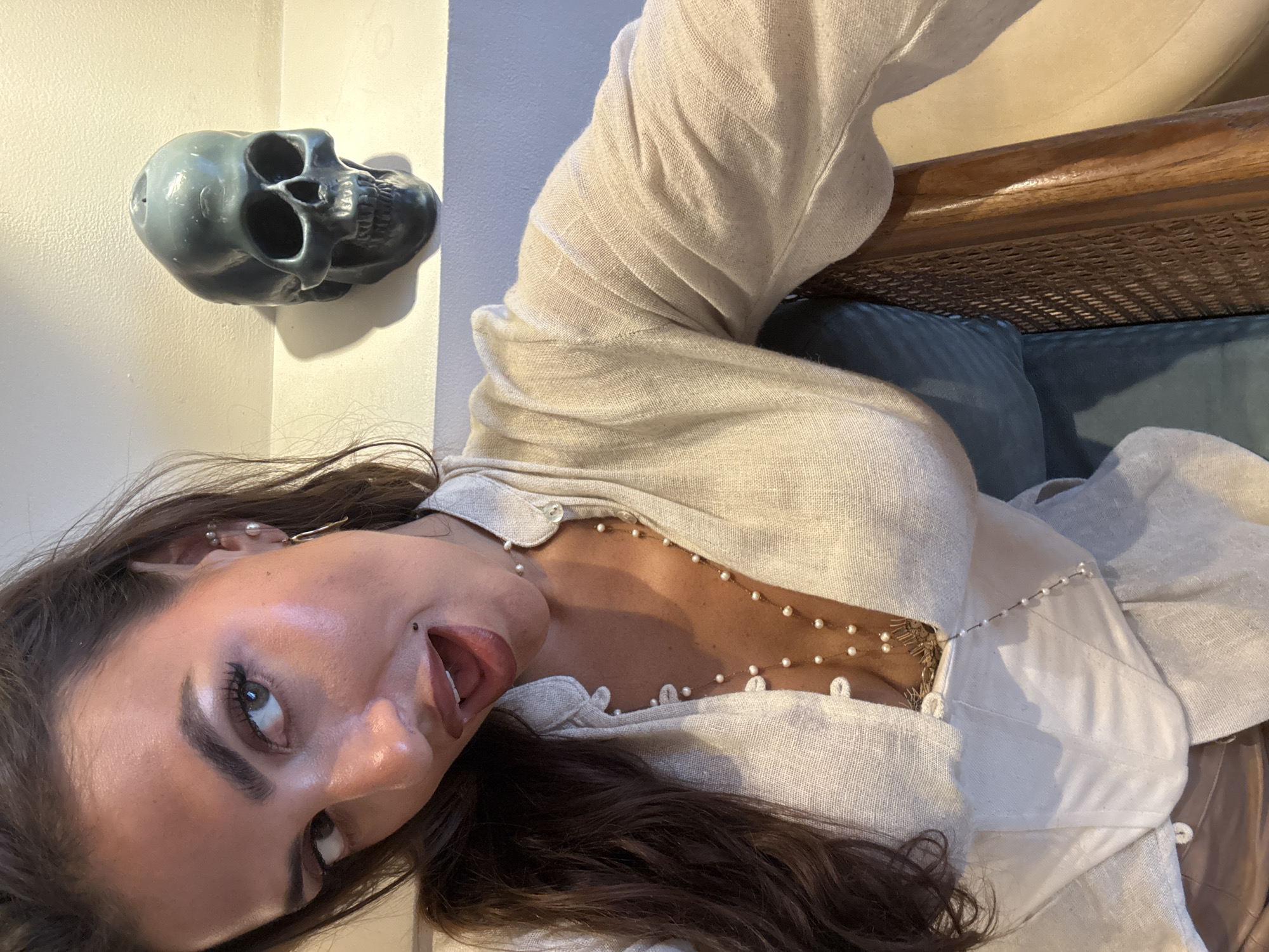 A woman is depicted in a medium shot, posed at an angle. She has dark hair and fair skin. She is wearing a light-colored linen blouse with pearl accents. Her mouth is slightly open. Behind her is a wall with a skull decoration. A wicker chair is also visible. The location is not discernible as North Kuta, Indonesia, based on the given image.