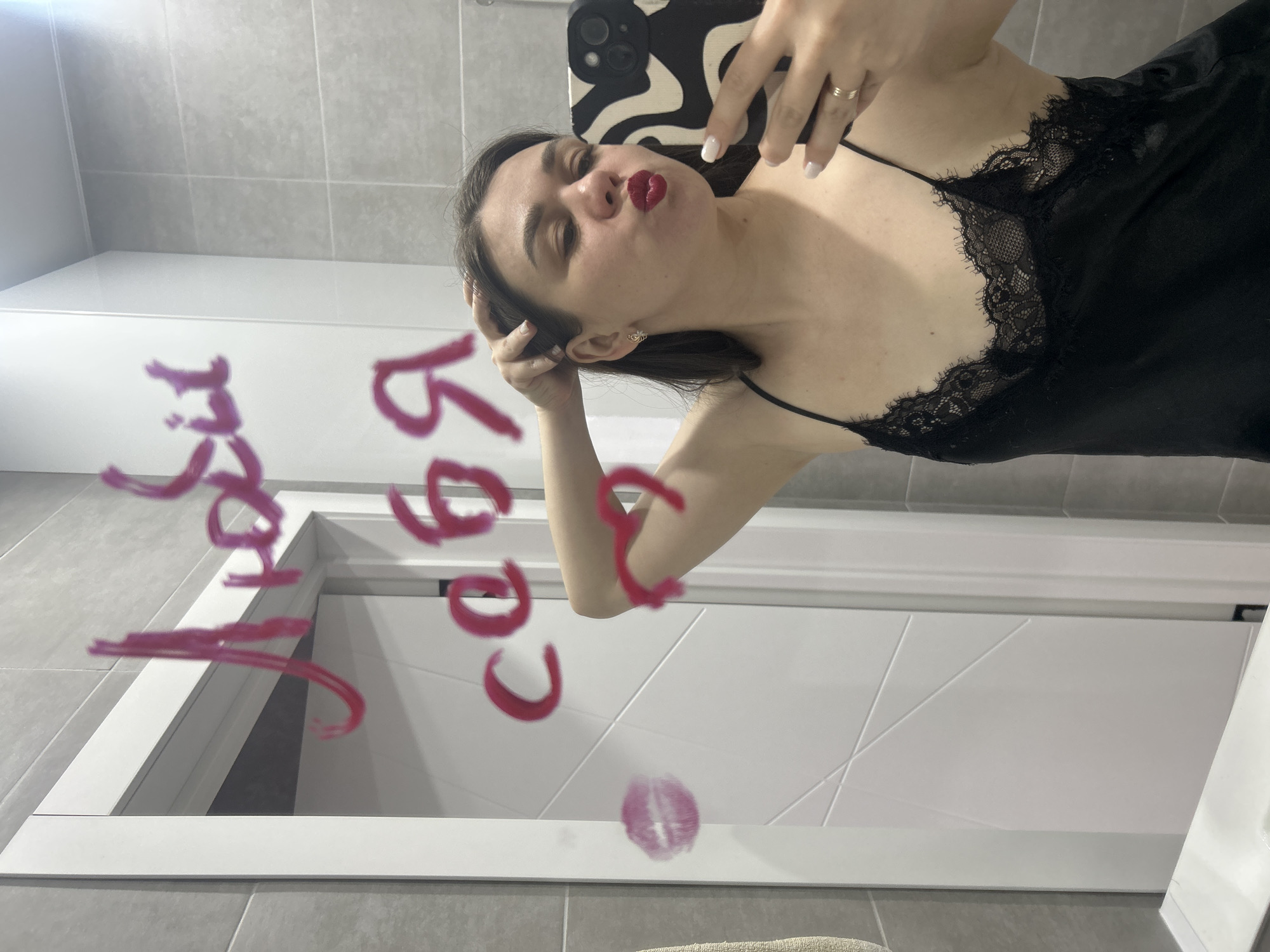 A young woman is captured in a rotated mirror selfie, striking a playful pose with pouted lips and vivid red lipstick. She has dark hair and is wearing a black, lace-trimmed slip dress or nightgown. Her left hand holds a smartphone with a distinct black and white patterned case, used to take the photo, while her right hand rests near her head. She displays white nail polish and a ring on her right ring finger.
The reflection in the mirror reveals a bathroom setting, characterized by light gray rectangular wall tiles and a white door with subtle geometric detailing. A white counter or shelf is also visible.
Prominently featured on the mirror, written in red lipstick, is Cyrillic text. Clearly visible are the words "любовь" (lyubov, meaning "love") and "себя" (sebya, meaning "self" or "yourself"), implying a message such as "love for oneself." Below the text, a distinct red lipstick kiss mark is left on the mirror's surface. The indoor scene is well-lit. The context of Chaikovsky, Russia, aligns with the Cyrillic script visible in the image.