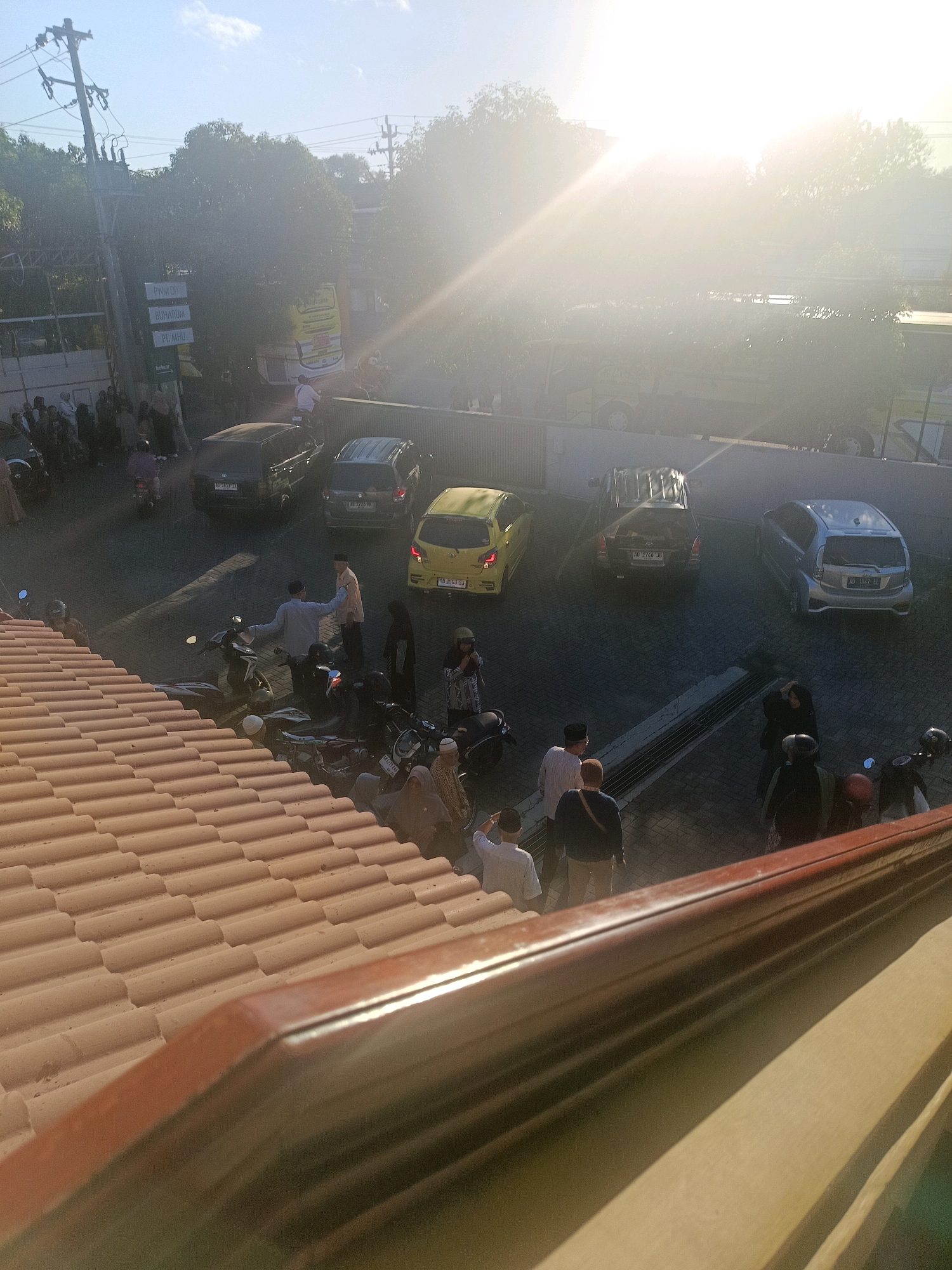 The image captures a sun-drenched scene in Yogyakarta, Indonesia, likely a street or parking area, characterized by a bright glare from the sun obscuring details in the upper portion. In the foreground, a tiled roof runs diagonally, casting a shadow on the area below. A street or paved area is visible, occupied by several parked cars, including a dark SUV, a silver hatchback, and a yellow car. Motorcycles are also present, some parked and others with people on or near them.
A group of individuals, predominantly dressed in traditional Islamic attire such as headscarves and robes, are scattered throughout the scene. Some appear to be conversing, while others are walking or standing. A few individuals are engaged in actions such as adjusting clothing or gesturing. The environment is partially obscured by trees, with utility poles and wires visible in the background. The lighting suggests it is either early morning or late afternoon, with the low angle of the sun creating strong highlights and shadows. The overall impression is one of everyday activity in a public space.