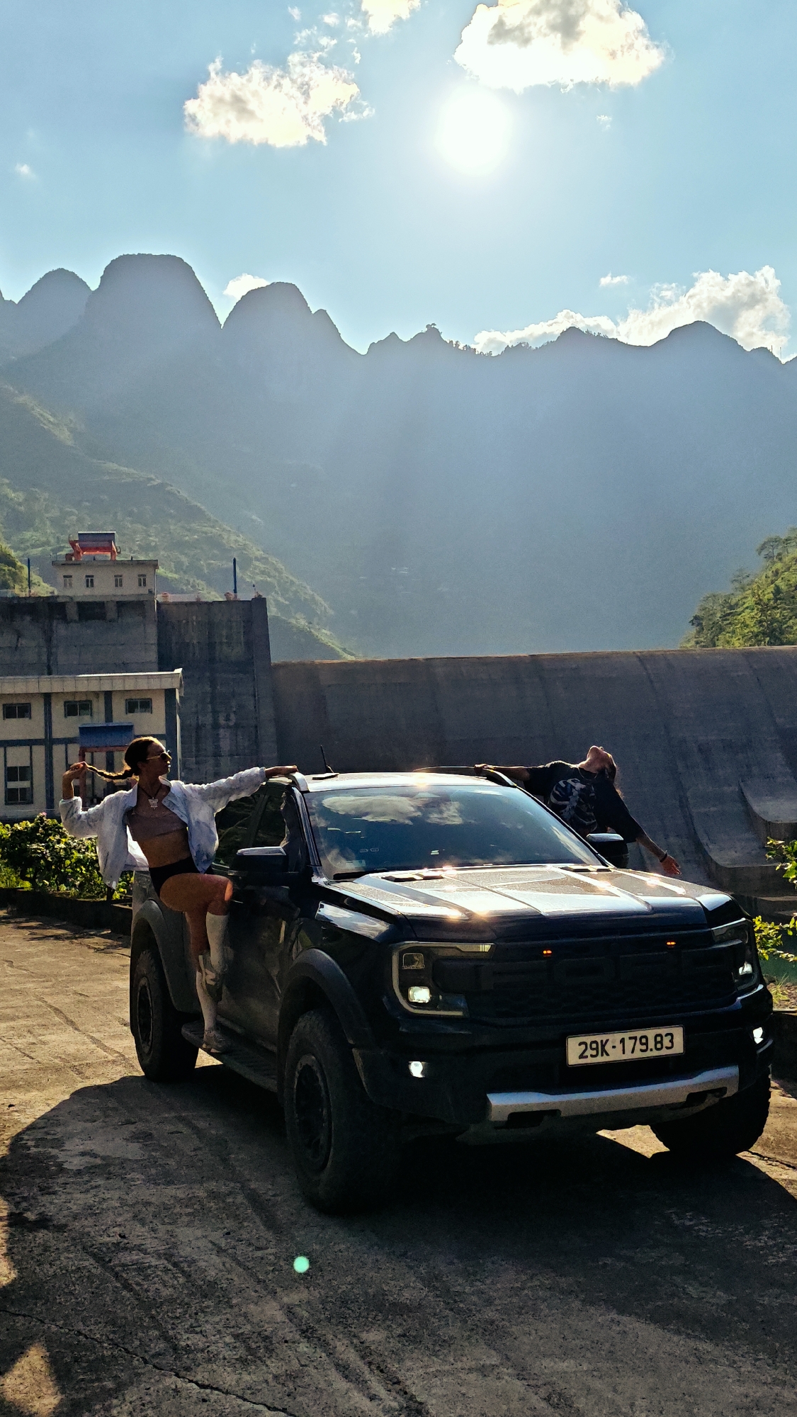 In Sơn Vĩ Commune, Vietnam, a black Ford Ranger with license plate "29K-179.83" is parked near a large concrete dam. A woman in a light jacket, top, and dark bottoms is perched on the driver's side, leaning on the roof. A man in a dark t-shirt is leaning over the passenger side of the vehicle. Behind the dam are forested mountains under a bright sky with scattered clouds. A two-story building is visible on the left, positioned against the dam's side. The ground is paved.