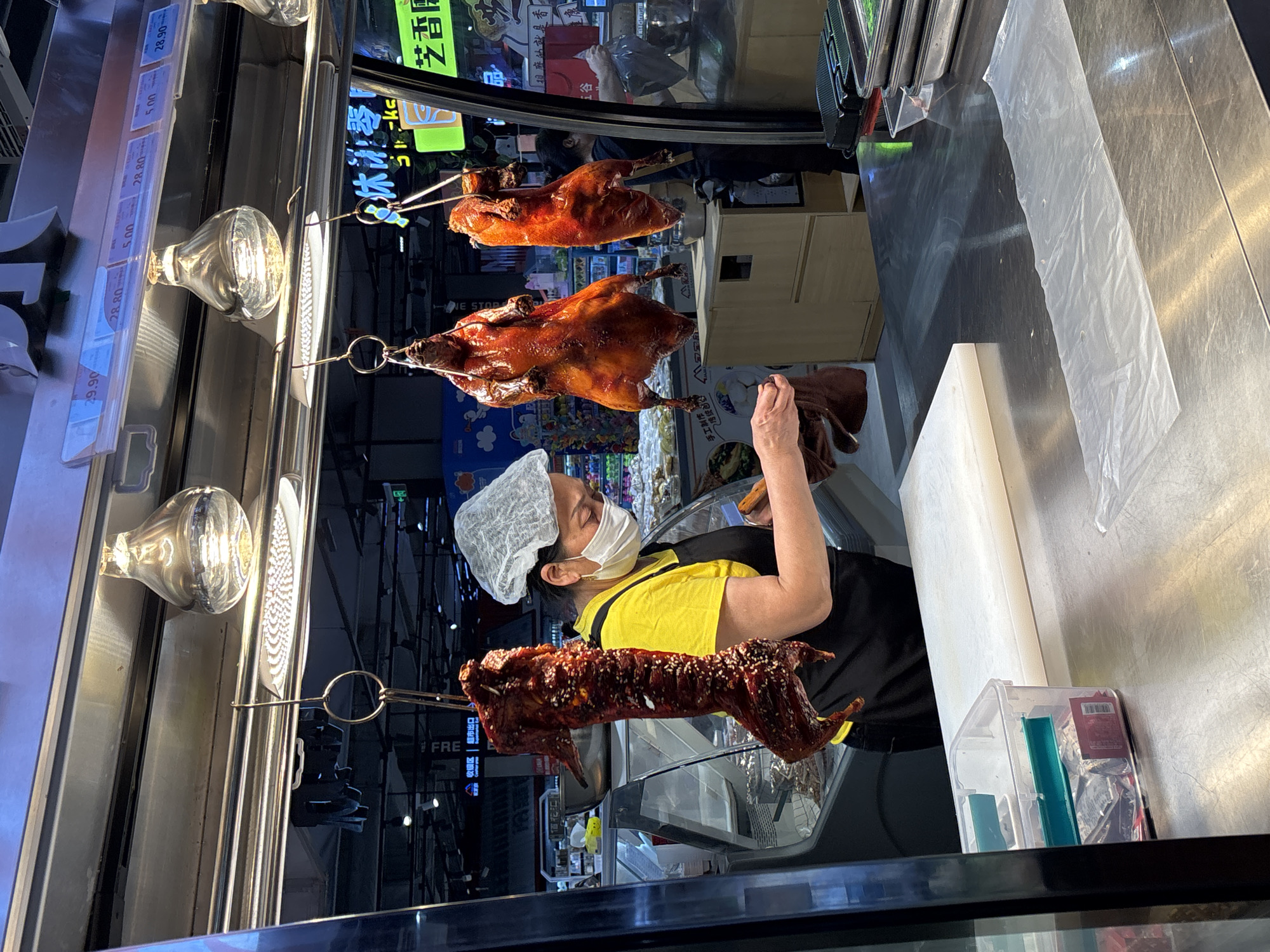 The image shows a food stall in Jiangjin, China. Three roasted ducks hang on metal hooks in front of a glass display case with a price list in Chinese. A woman in a white hairnet and face mask, wearing a yellow shirt and black apron, is visible behind the display case. The stall is located inside of a grocery store.
