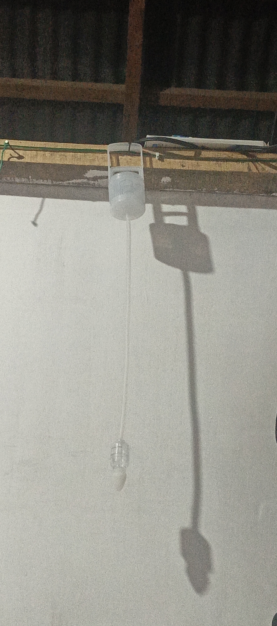 A clear plastic water bottle with a string attached to its cap hangs from a wooden beam. A thin, clear plastic tube extends from the bottle and leads down to a small, clear plastic enclosure at its end. The scene appears to be indoors, with a corrugated metal roof visible in the background. A shadow of the hanging bottle and tube is cast on a white wall. There are no people or animals present. This scene is located in Cinta Dame, Indonesia.