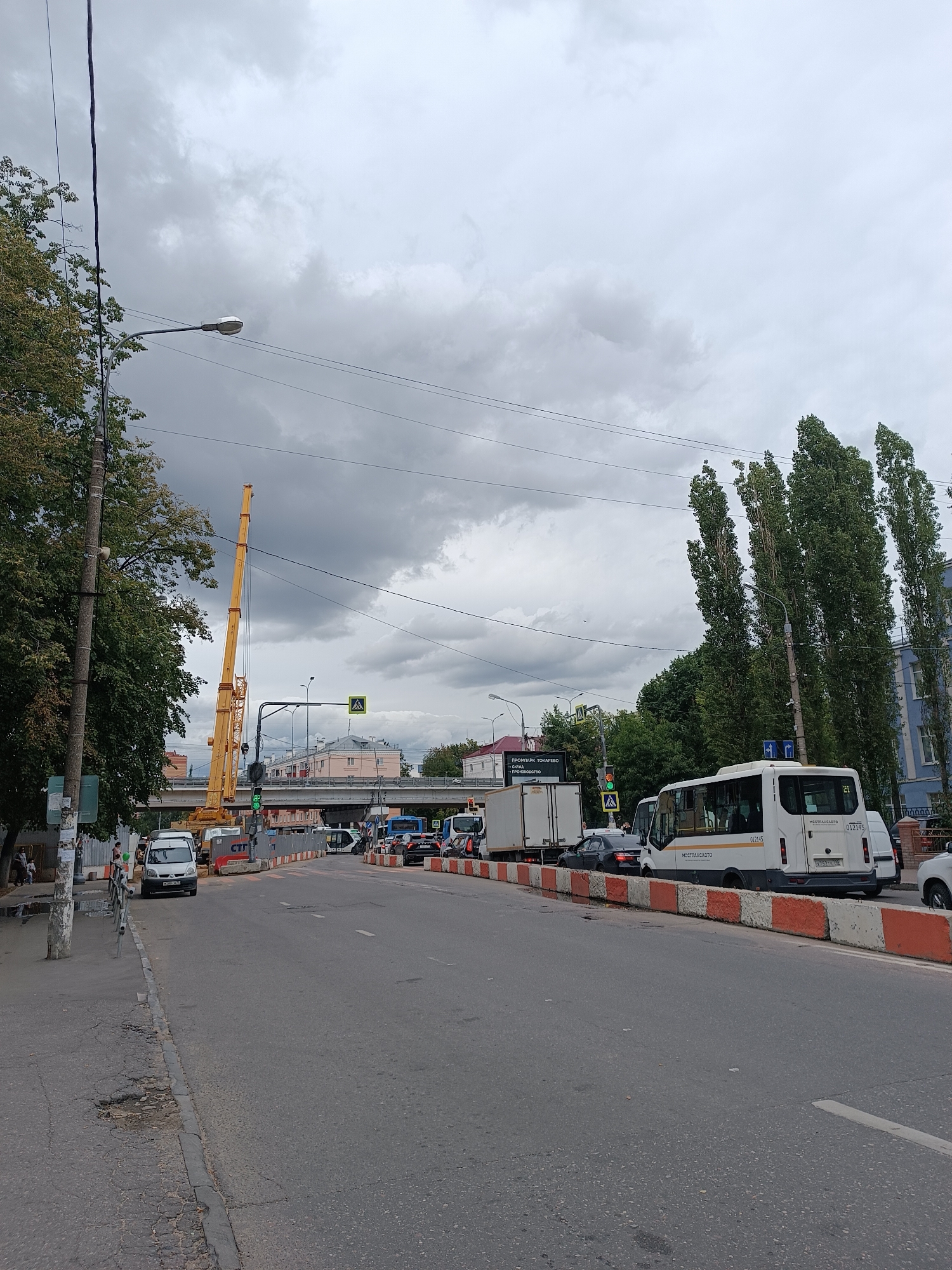 The image depicts a street scene in Lyubertsy, Russia, on a cloudy day. The foreground shows an asphalt road with some cracks and a worn curb. Orange and white concrete barriers line the right side of the road, separating it from what appears to be a construction area or closed-off section. In the middle ground, several vehicles are visible, including a white bus with "МОСТРАНСАВТО" branding, a white truck, and several cars. A yellow crane is prominently positioned on the left side of the road, suggesting ongoing construction. Traffic lights and pedestrian crossing signs are also present. The background features buildings, trees, and an overhead bridge or overpass. Power lines stretch across the sky, which is filled with grey clouds. A few pedestrians can be seen on the left sidewalk, near a lamppost and some trees.