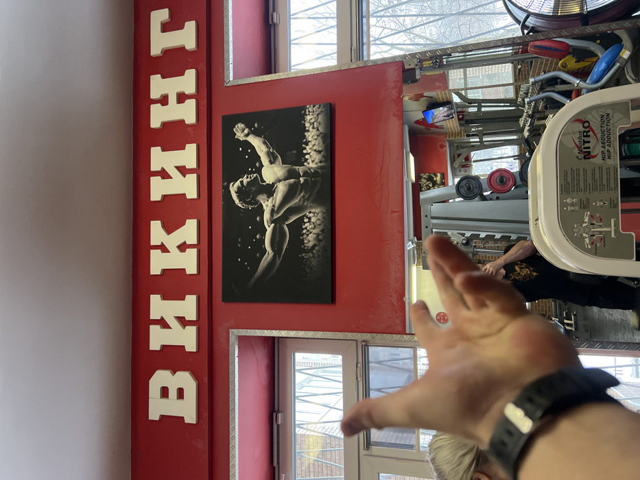 Inside a gym, a red wall displays white Cyrillic letters "ВИКИНГ" vertically. A black and white muscular bodybuilder poster hangs next to it. Gym equipment is visible, including weight machines. A person extends their hand towards the camera, wearing a black wristwatch. A window with a metal frame is partially visible behind their hand.