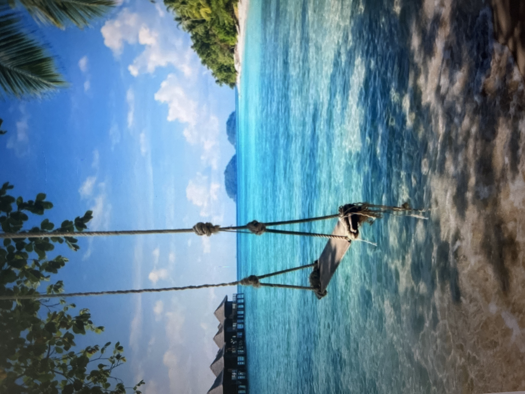 A rope swing hangs over the clear, turquoise water of a tropical beach. The swing, made of weathered wood and thick rope, is suspended from branches of a leafy tree in the foreground. In the mid-ground, a row of thatched-roof overwater bungalows are visible in the calm sea, suggesting a resort location. Further in the distance, lush green islands dot the horizon under a bright blue sky with scattered white clouds. The shoreline is lined with palm trees, and the sandy beach is visible where the water meets the land. Sunlight reflects off the water, creating dappled patterns of light and shadow on the seabed.
