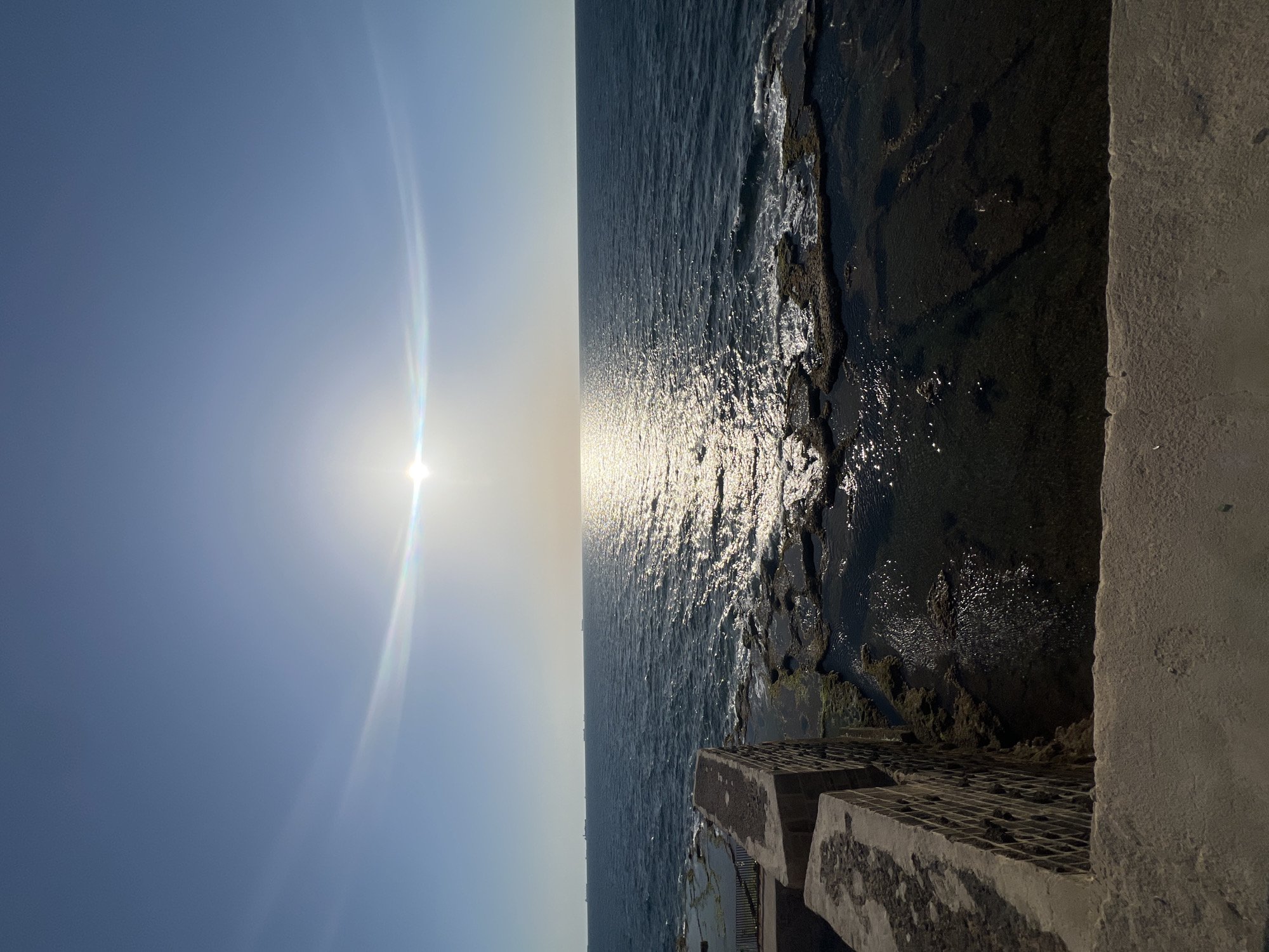 The image captures a vibrant seascape under a clear sky, with the sun low on the horizon. The horizon line stretches horizontally across the middle of the frame, dividing the sky and the sea.
The bright sun, positioned on the left side of the frame, creates a dazzling path of light reflecting off the water's surface, extending towards the viewer. This glare creates a sparkling effect on the sea, which appears dark blue to grey in other areas, indicating depth. Small waves are visible, especially closer to the shore. The sky is a gradient of light blue, becoming almost white near the sun and the horizon, with visible lens flare emanating from the sun.
Along the right side of the image, a dark, rugged coastline or rocky shore meets the water. In the foreground on the bottom right, large, grey concrete structures are visible. These appear to be sturdy blocks, possibly part of a seawall, pier, or breakwater, with some showing a textured or grid-like surface on top. The water around these structures appears shallower and darker, with some green growth possibly visible on the submerged or wet rock surfaces.
The scene depicts a serene and bright moment, likely during late afternoon or early morning, showcasing the interaction of sunlight with the sea and coastal structures in a location such as Acre, Israel. There are no people visible, and no discernible text.
