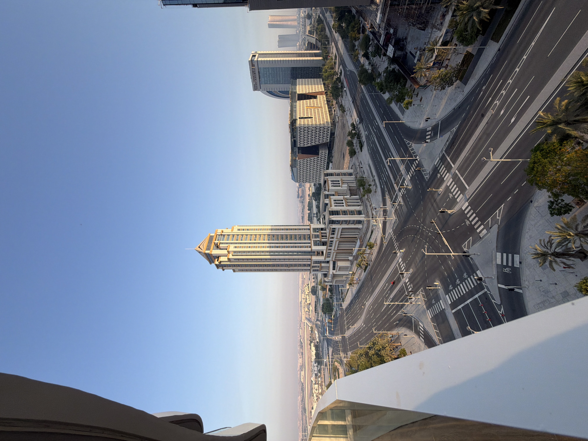 This is an aerial, high-angle, eye-level shot of a city intersection and surrounding buildings at dawn. The sky is a clear, pale blue with a slight haze near the horizon. A contemporary, tall, rectangular skyscraper with a golden-colored pyramidal roof dominates the center-right of the image. It features a grid of windows and decorative pillars at its base. To its left, a large, angular building with a patterned facade is visible, and further back, more modern skyscrapers of varying shapes and sizes can be seen stretching into the distance. An empty, multi-lane intersection with white road markings and pedestrian crossings is the focal point of the foreground. Streetlights and palm trees line the roads. A white balcony railing occupies the far left of the frame, suggesting the photo was taken from a high vantage point within a building. The scene is quiet and devoid of significant traffic or pedestrian activity, typical of early morning hours.