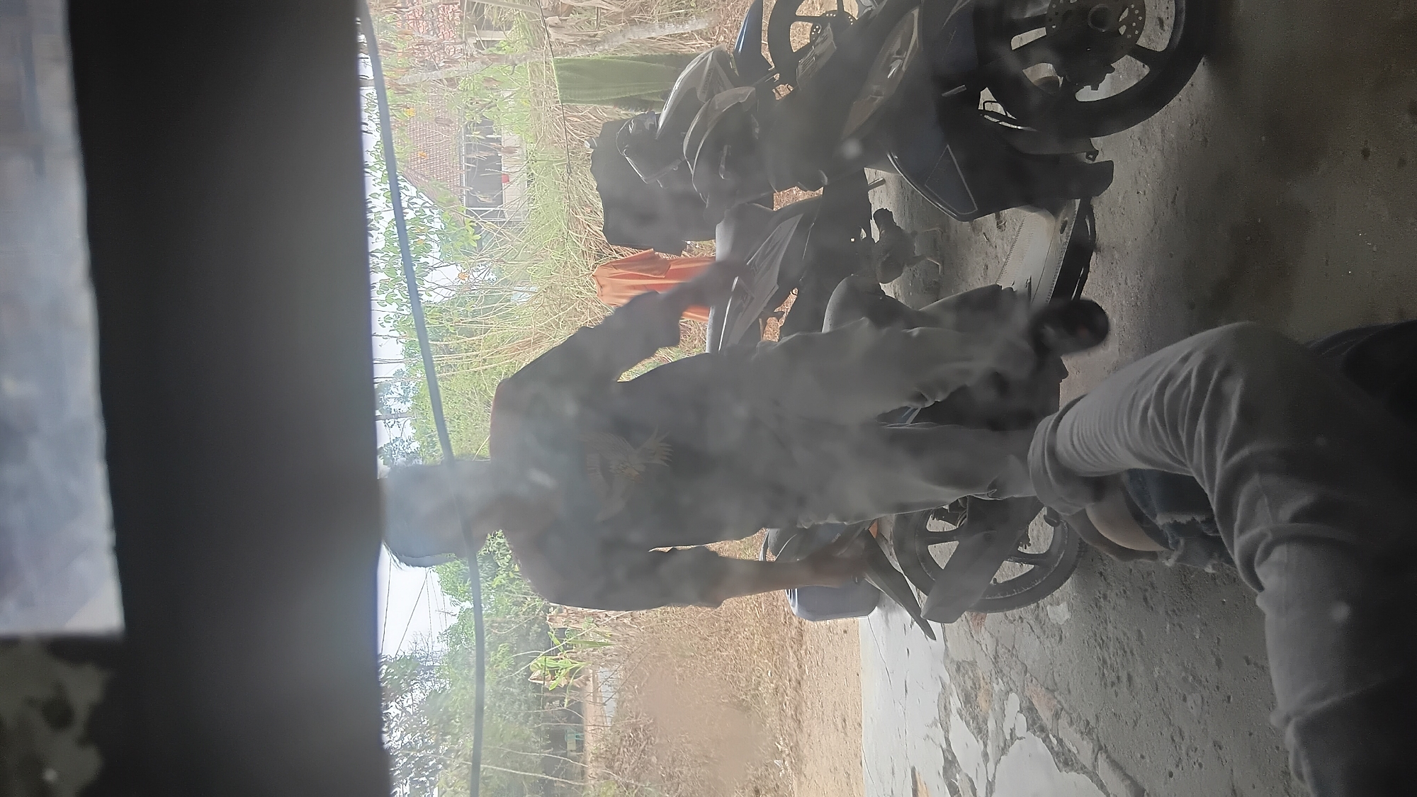 The image depicts an outdoor scene, possibly in Prabumulih, Indonesia, characterized by a slightly dusty and sunlit environment. In the foreground, a person's arm and shoulder are visible, clad in a grey long-sleeved shirt, reaching towards the lower right. The main subject is a person standing next to a dark-colored scooter, also wearing a grey long-sleeved shirt with a graphic on it. This individual appears to be in the process of interacting with the scooter. Behind them, another scooter is partially visible. In the background, there are trees and vegetation, along with a rustic building. A piece of orange fabric and some green fabric are hanging, possibly laundry. A small chicken is visible on the ground near the scooters. The overall impression is of a casual, everyday scene, possibly a residential yard or a workshop.
